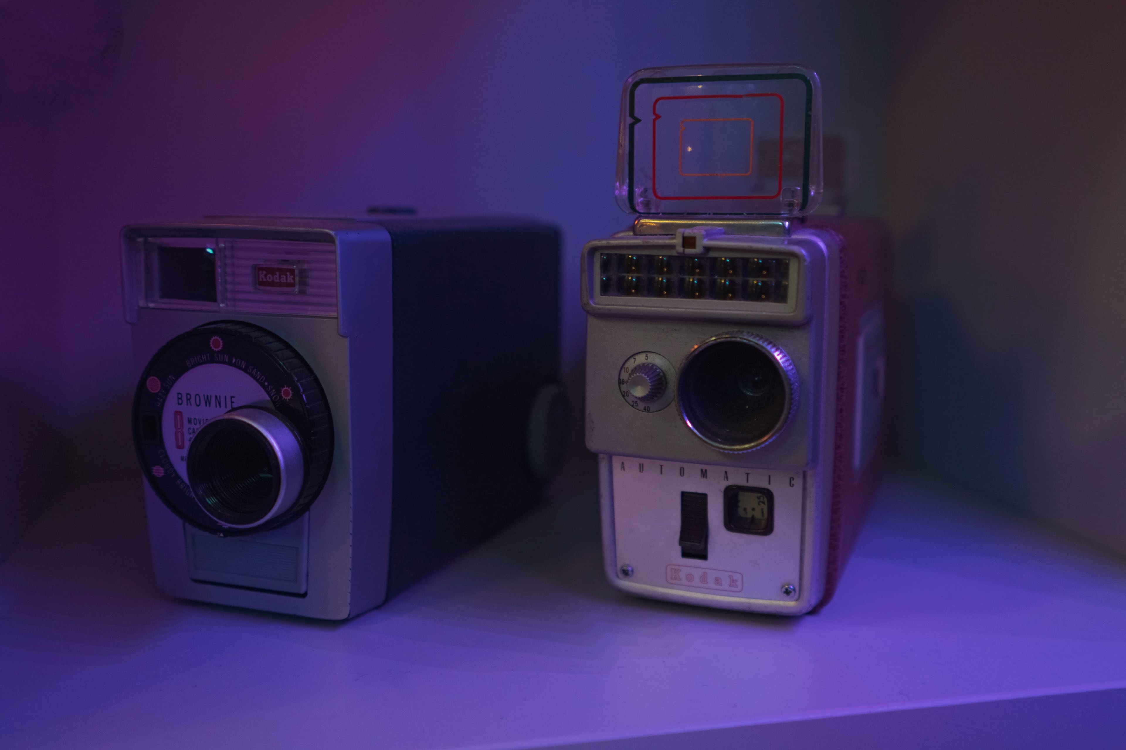 Two vintage Kodak cameras displayed on a shelf, illuminated by purple lighting.