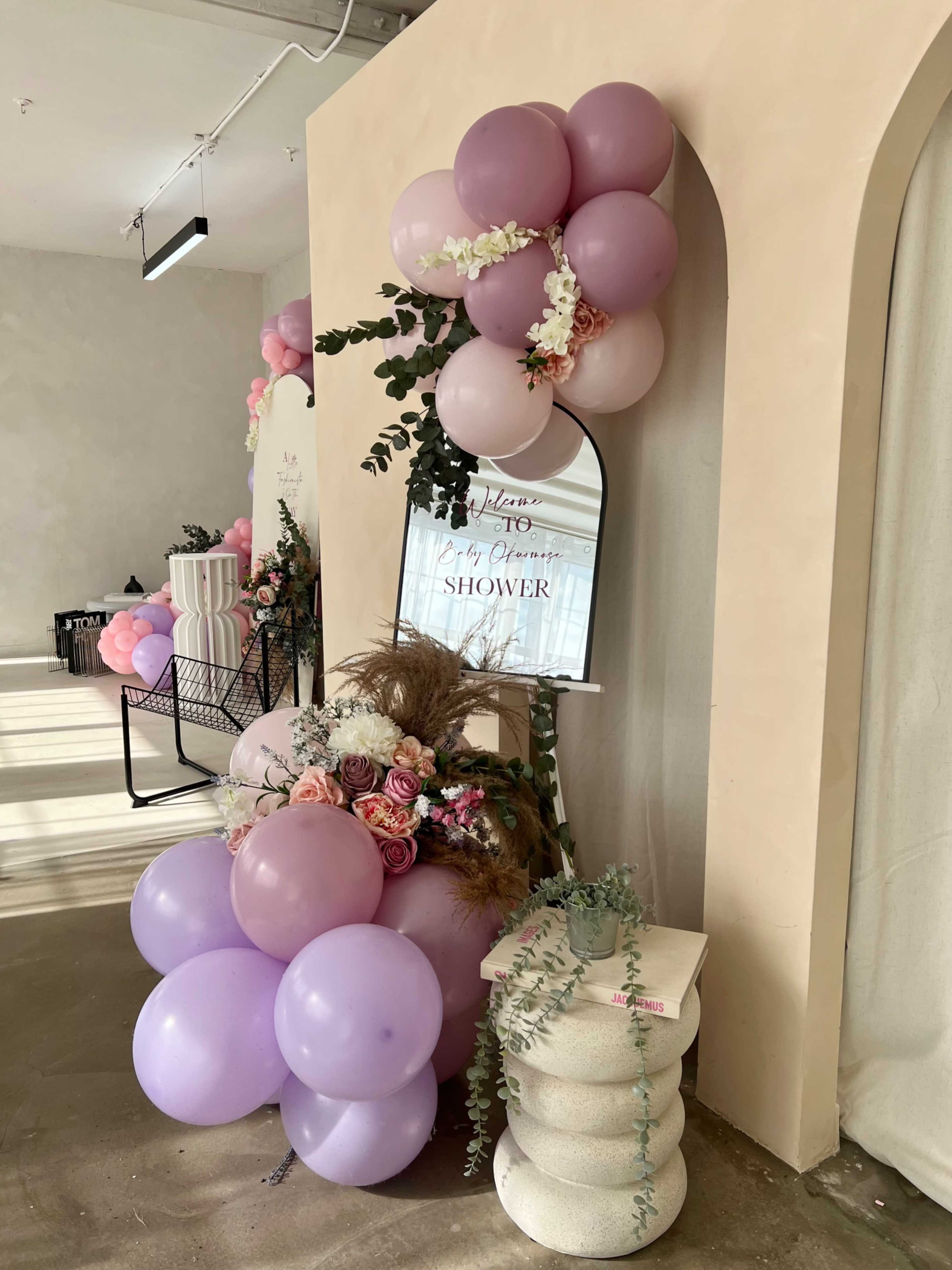 The image depicts a decorative display for a baby shower, featuring clusters of pink and purple balloons, flowers, and a mirror along with a sign that reads "Baby Shower."