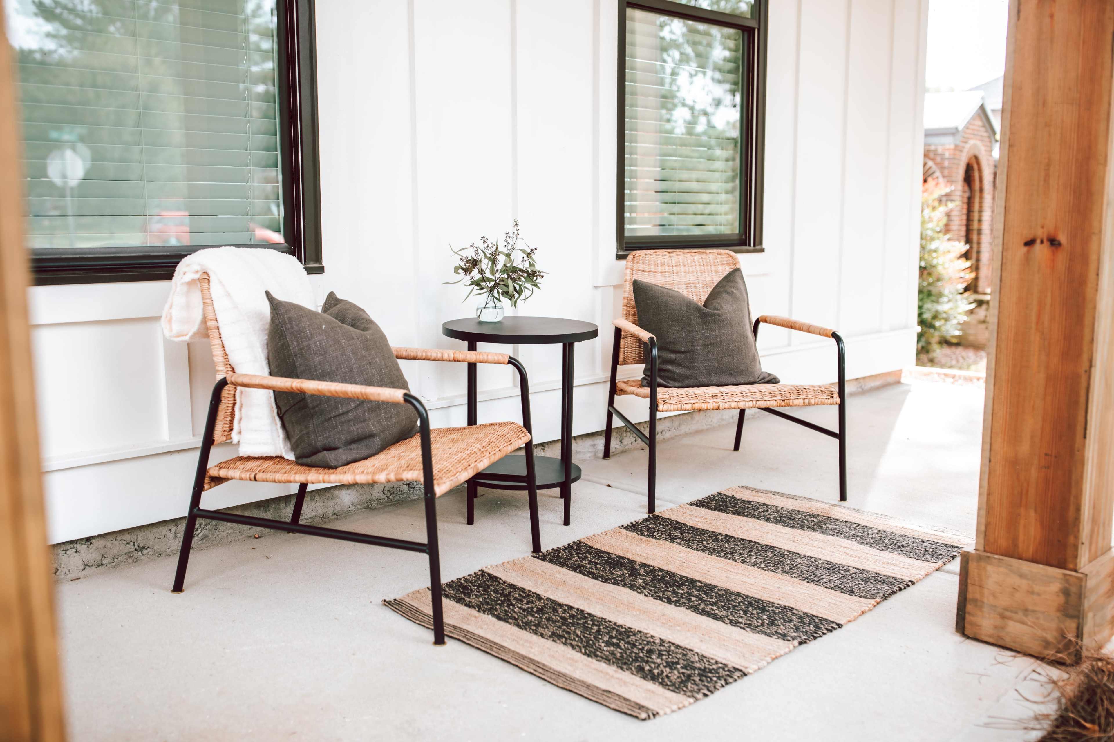 The image shows a cozy porch area featuring two chairs with cushions, a small round table, and a striped rug on a concrete floor.