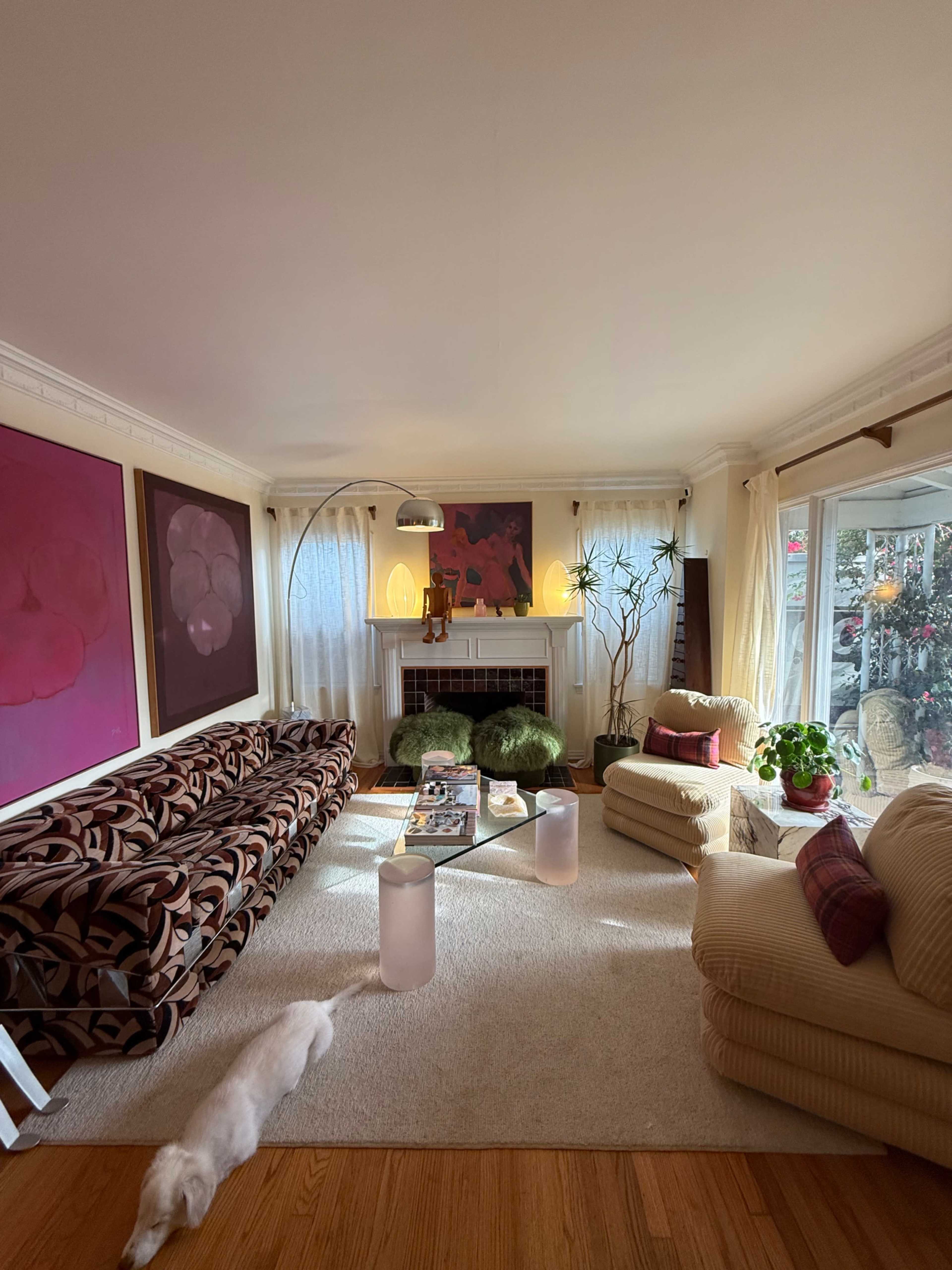 The living room features a patterned sofa, a beige armchair, a coffee table with magazines, and large windows allowing natural light to illuminate the space.