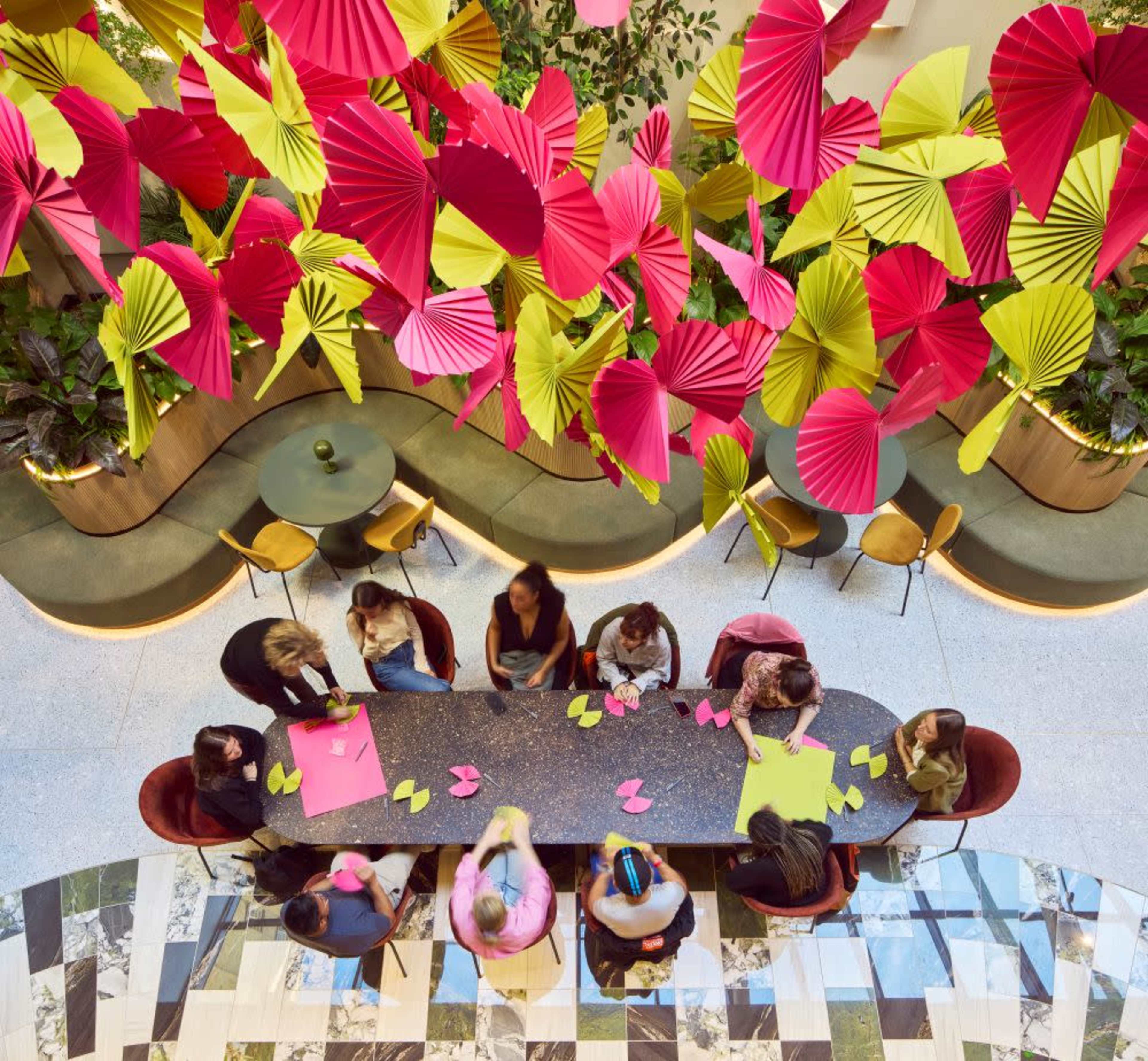 A group of people is sitting around a long table in a brightly decorated space with colorful, oversized paper decorations hanging overhead.