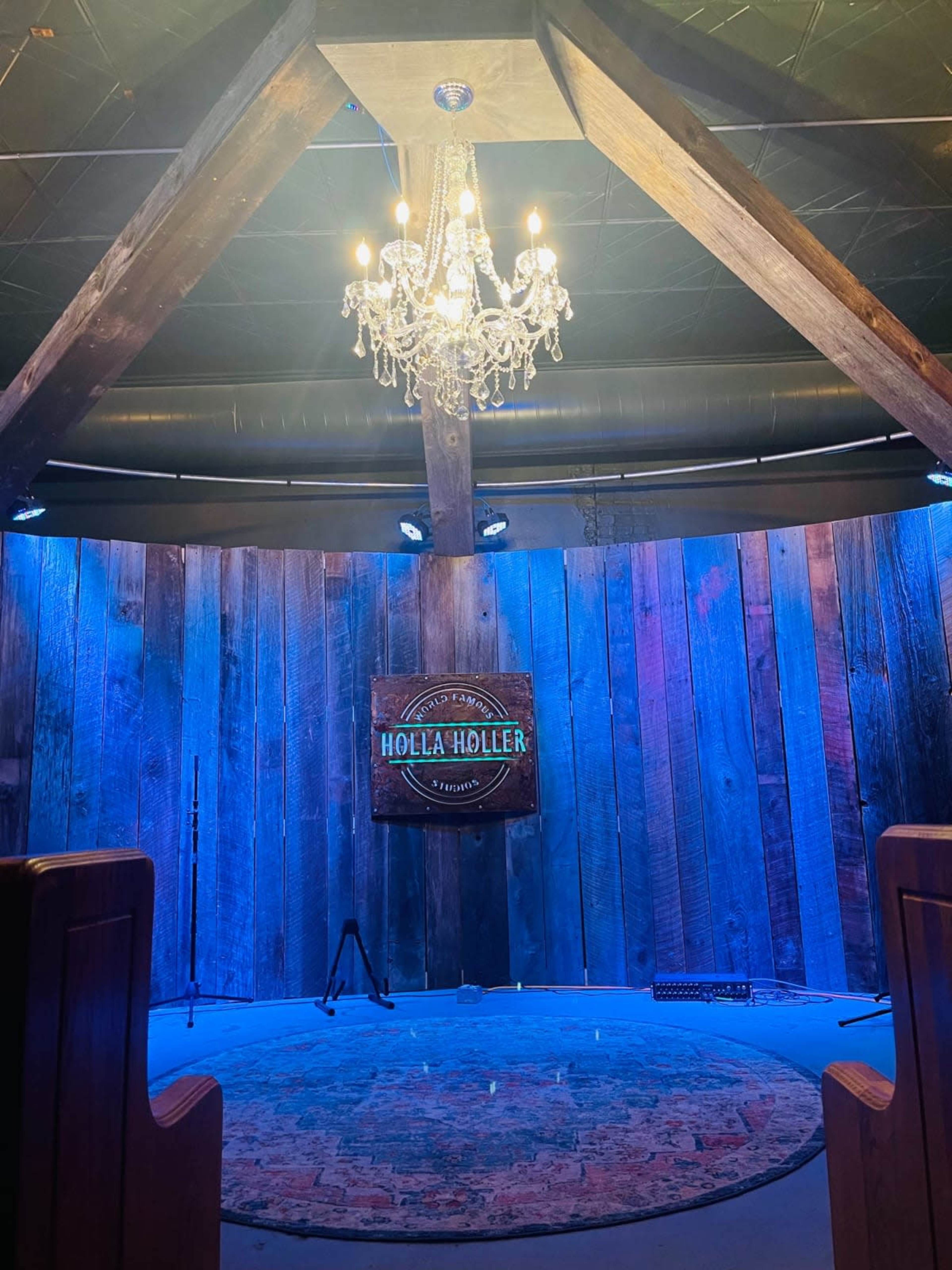The image shows an indoor performance space featuring a wooden circular stage, a chandelier hanging from the ceiling, and blue lighting illuminating the wooden walls.