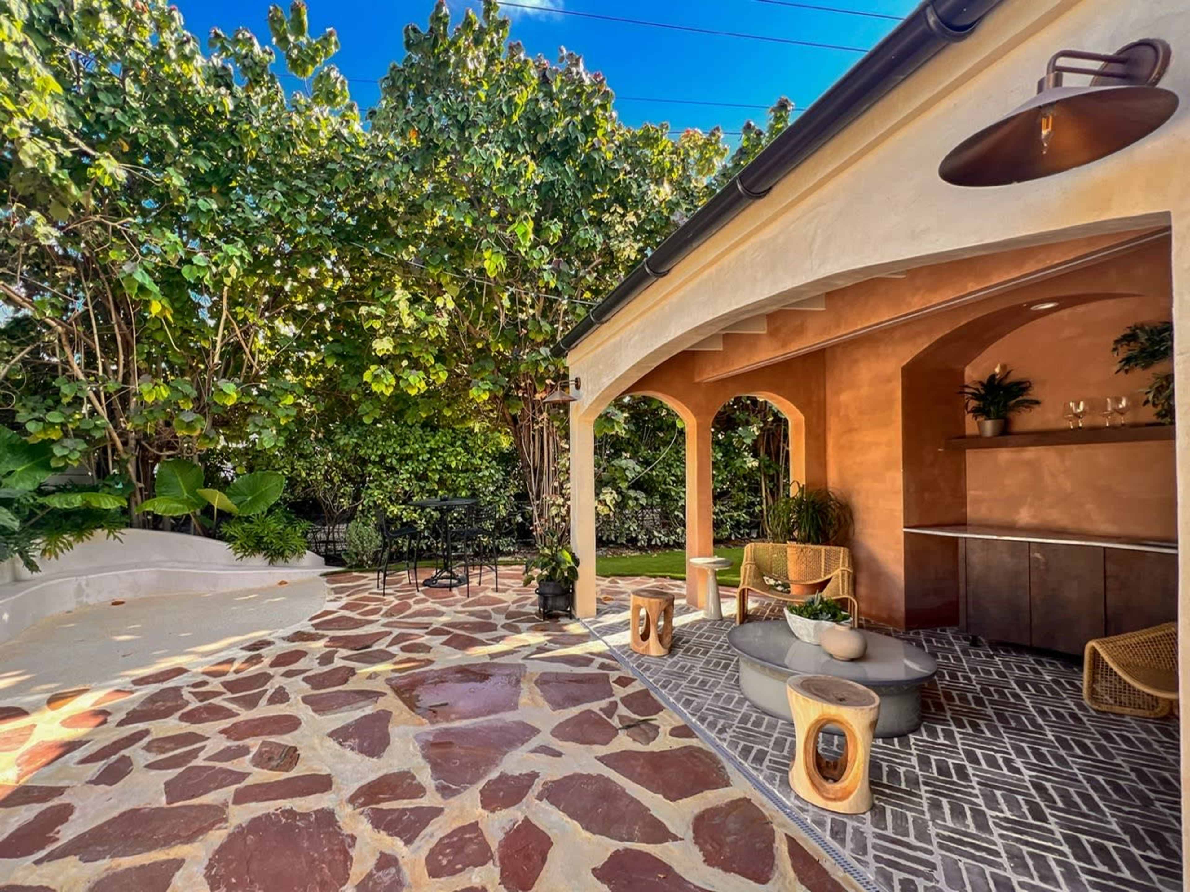 The image features a landscaped outdoor area with a stone patio, surrounded by greenery and a bar-style seating area.