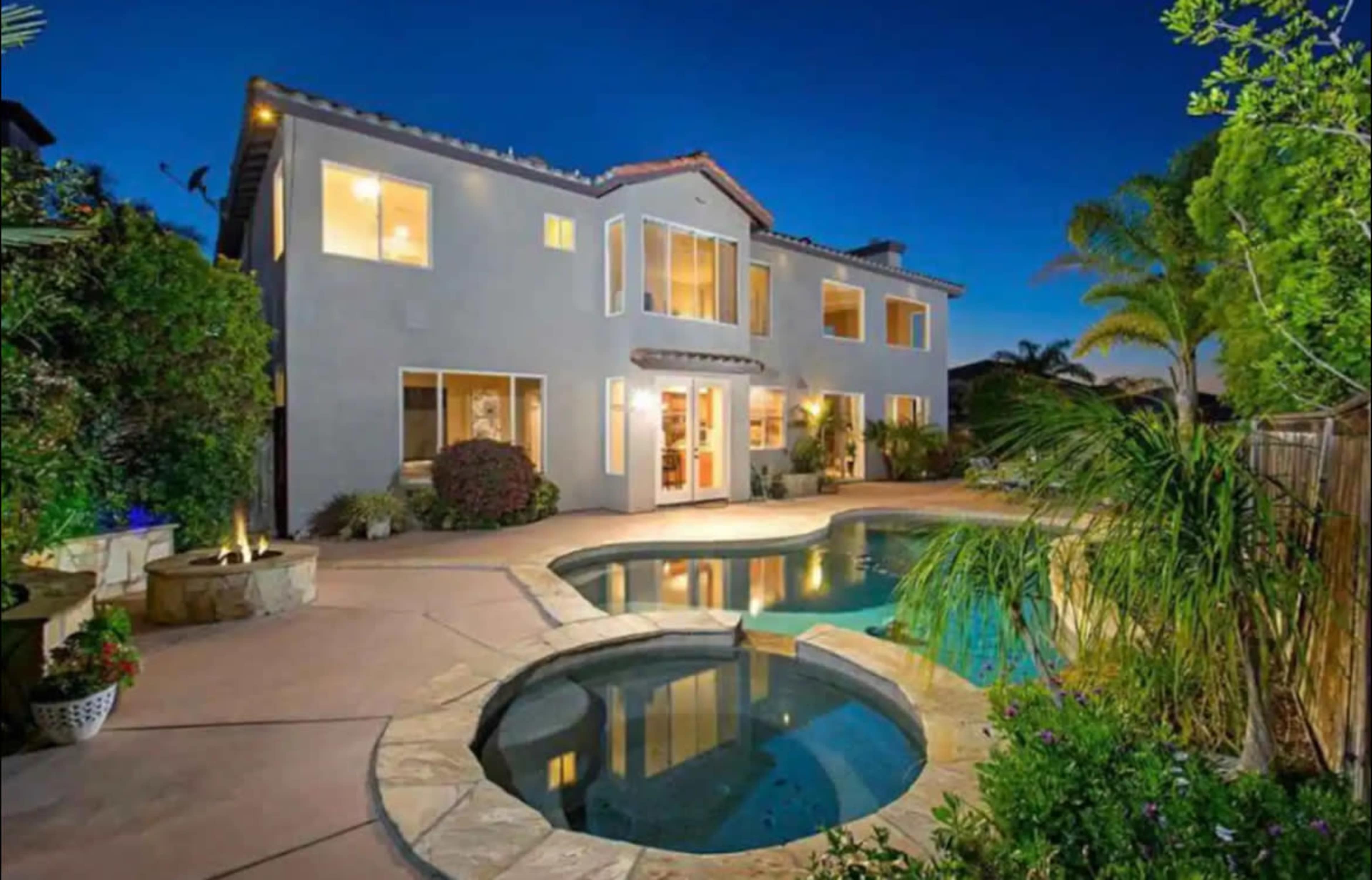 The image shows a two-story house with large windows and a swimming pool in the backyard, surrounded by palm trees and landscaping at dusk.