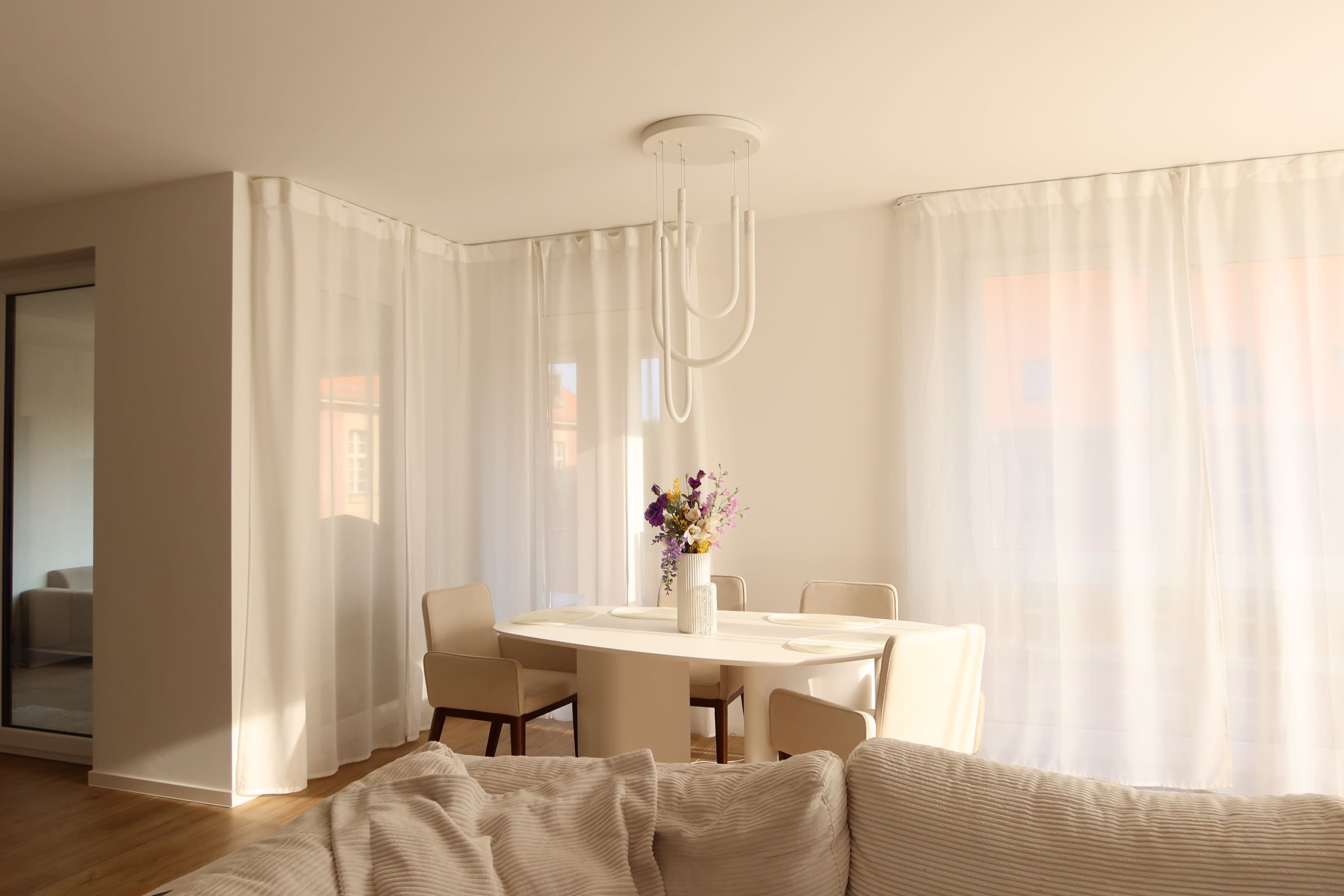 A minimalistic dining area features a white table surrounded by four chairs, illuminated by a modern ceiling light, with sheer curtains softly filtering natural light.