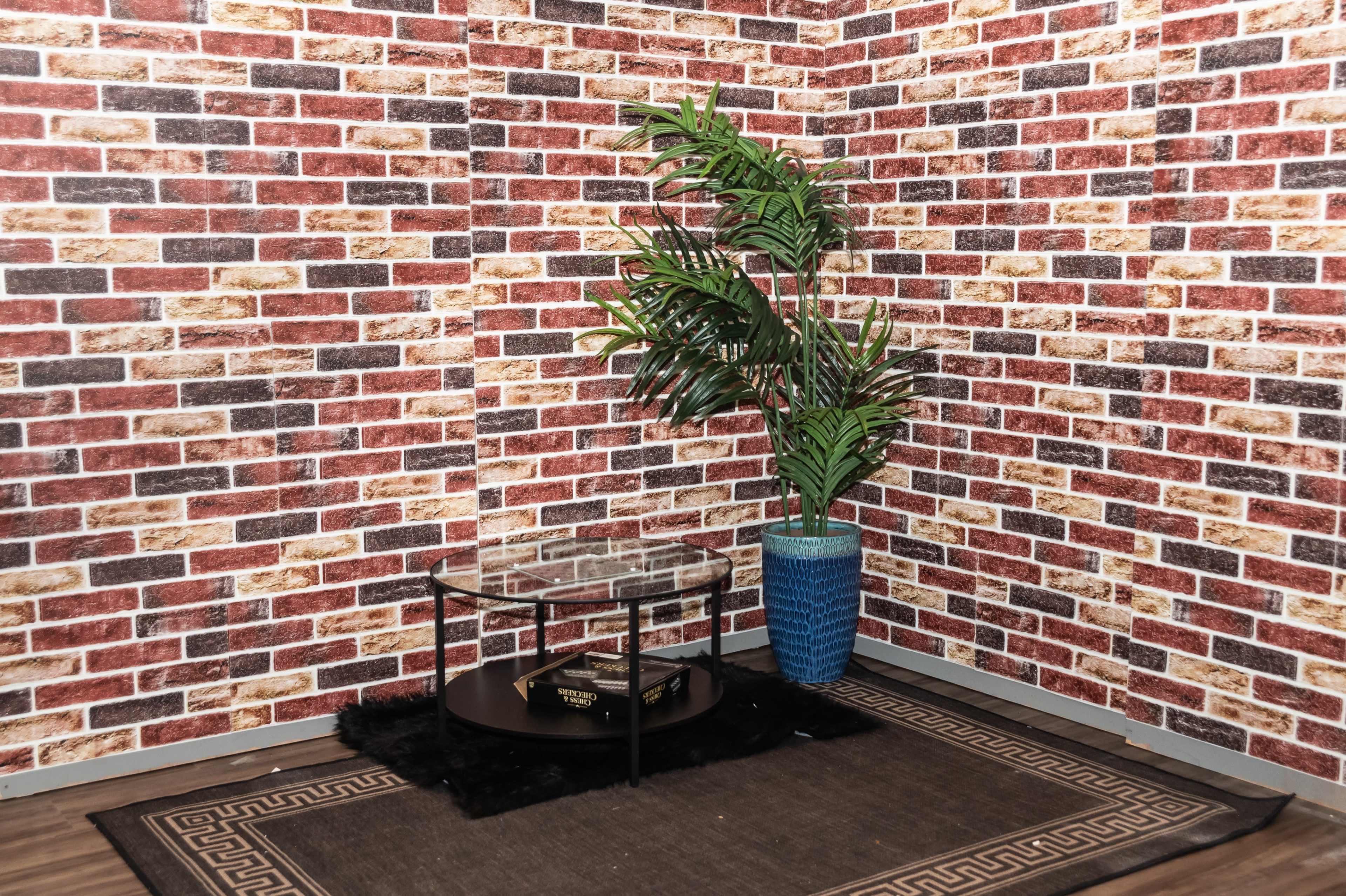 A corner of a room features a glass-top table on a black rug, surrounded by brick-patterned wallpaper and a green plant in a blue pot.