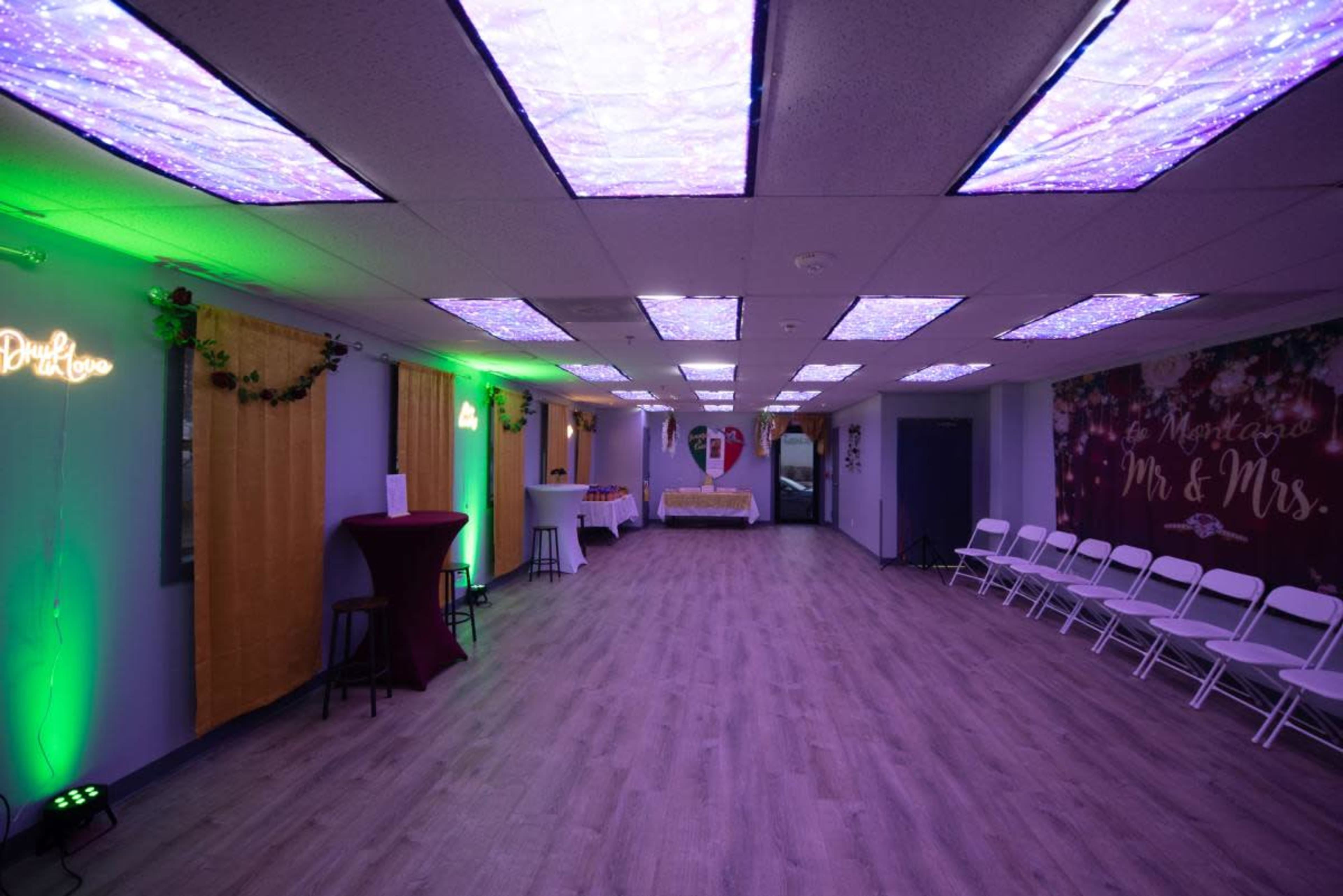 The interior of a decorated event space features purple and green lighting, a table with a lace tablecloth at the end, and rows of white chairs set up for guests.