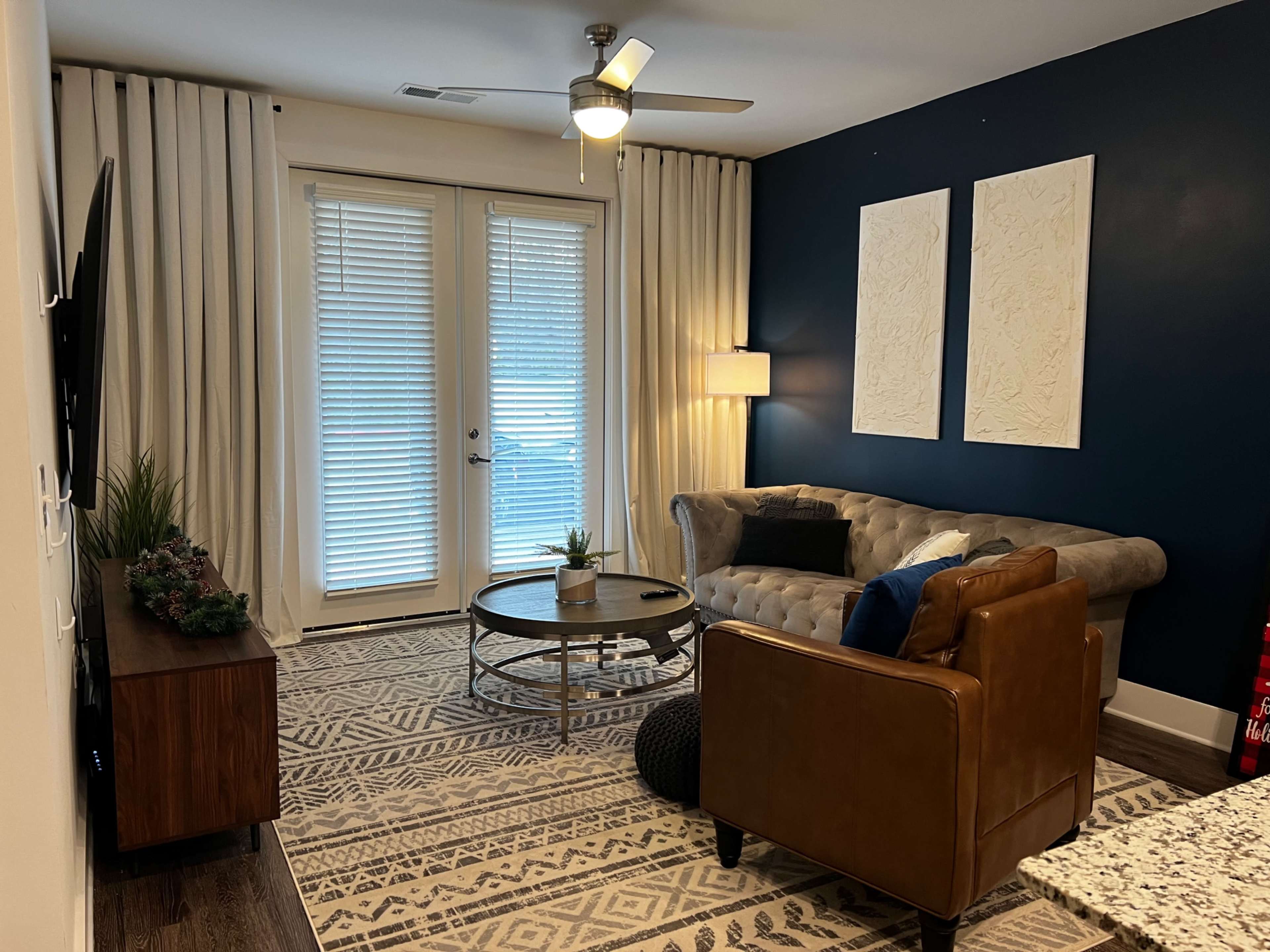A modern living room features a tufted sofa, a round coffee table, and a wall with two decorative panels, set against a dark blue backdrop and illuminated by natural light from sliding glass doors.