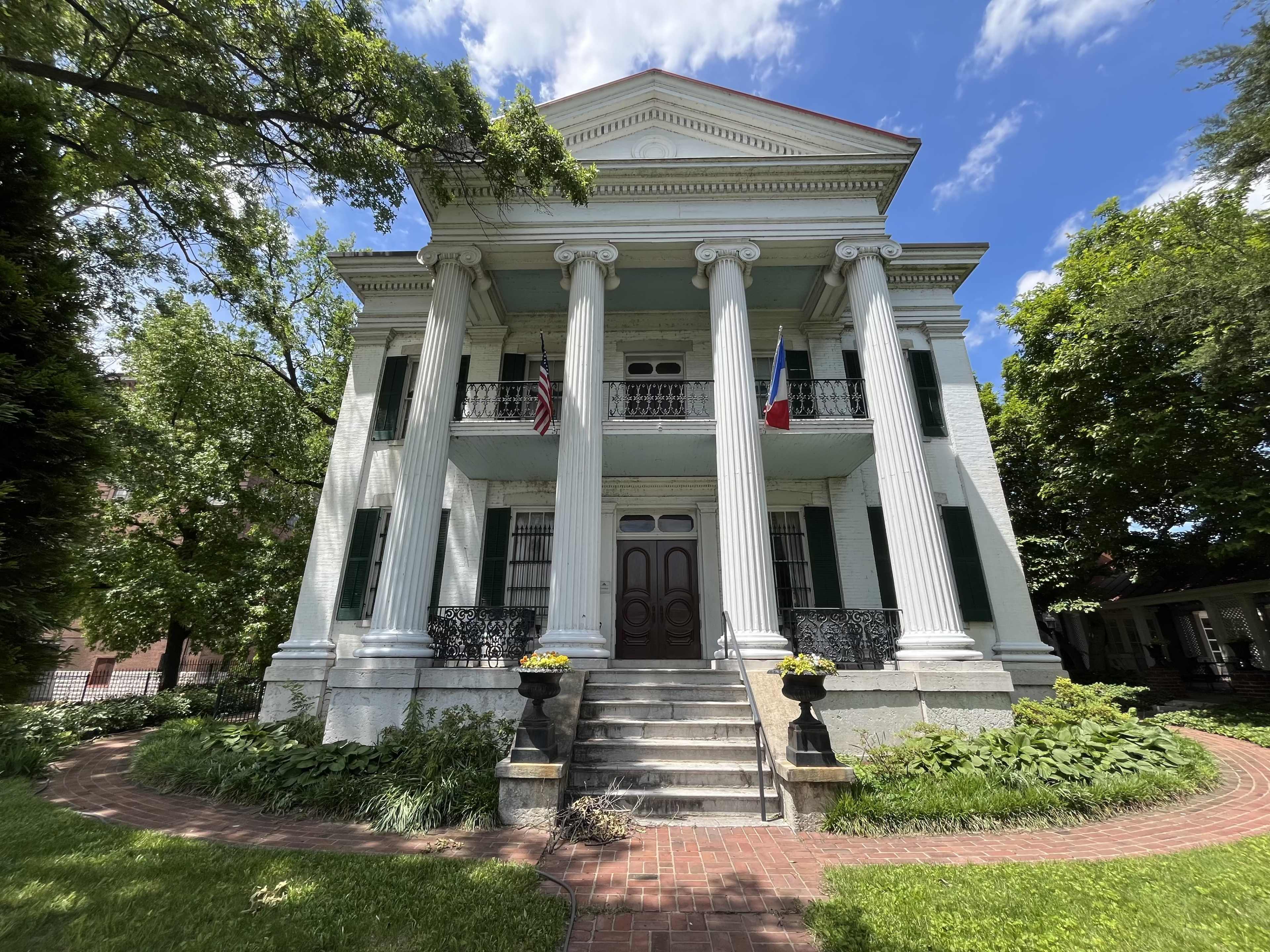 The image shows a historic two-story brick house with four large columns, two balconies, and lush greenery surrounding the front steps.
