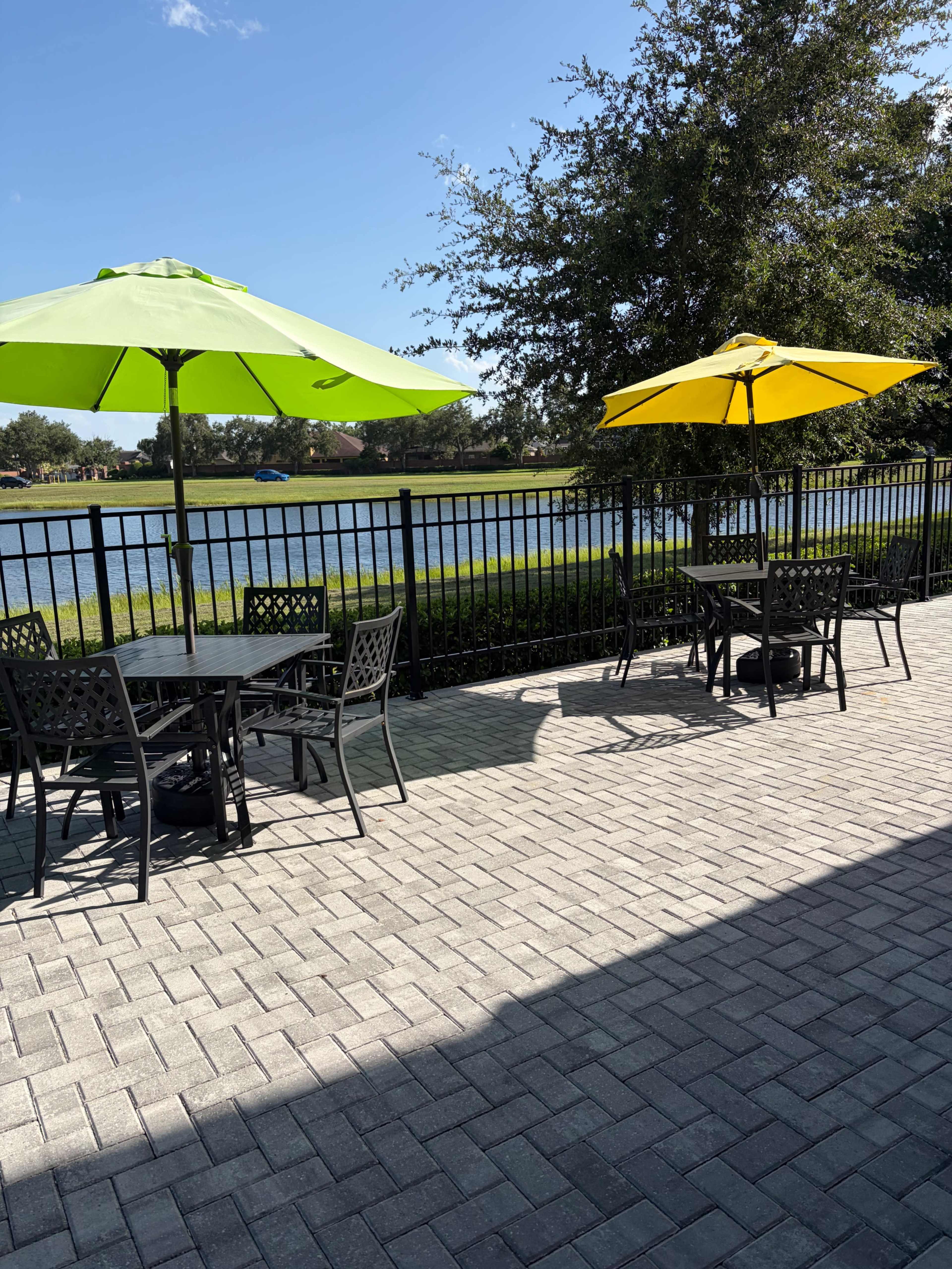 The image shows a patio area with black metal tables and chairs beneath green and yellow umbrellas, adjacent to a calm body of water.