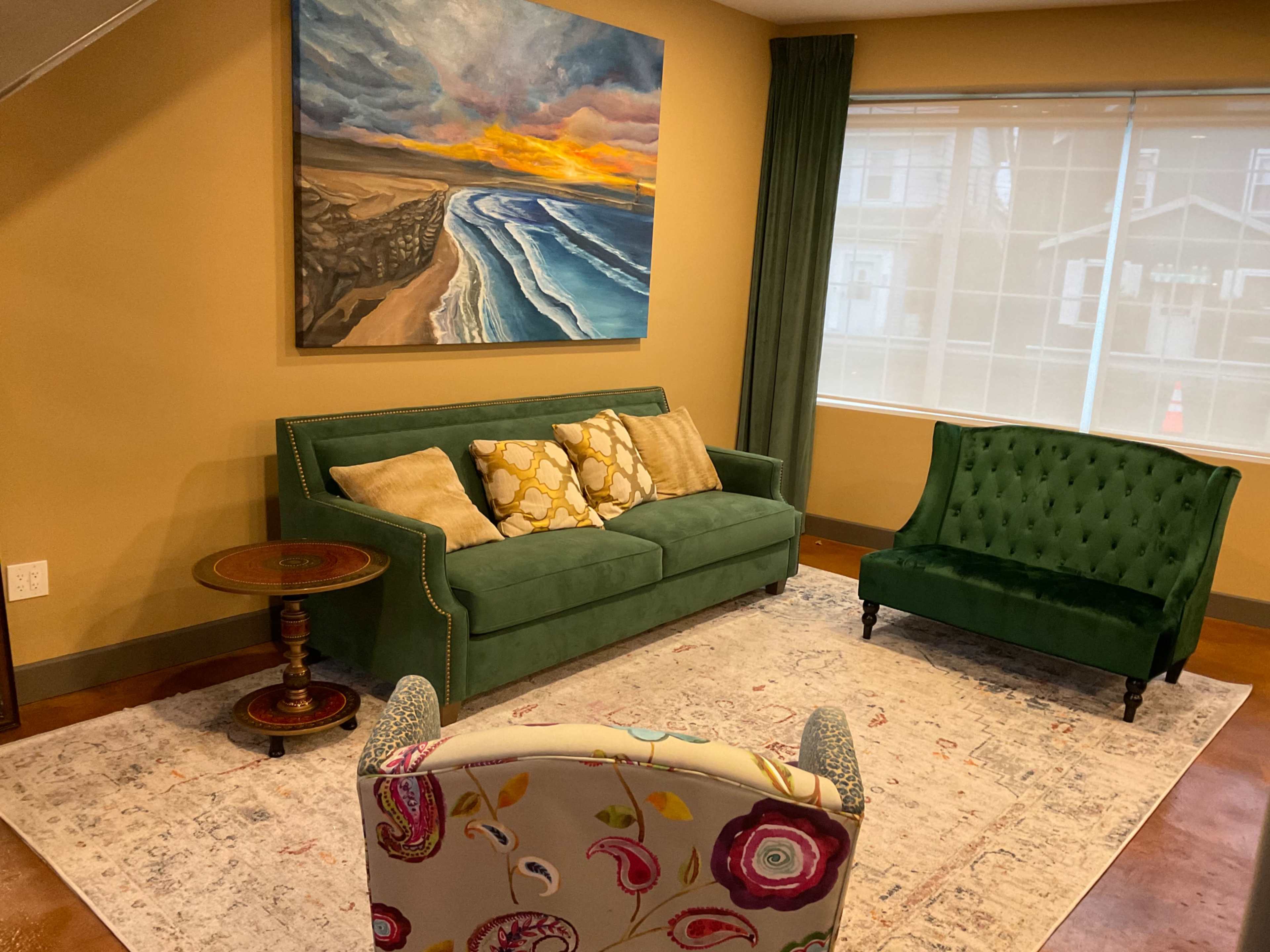 A cozy living room featuring a green sofa with patterned cushions, a green armchair, a small circular table, and a large painting of a beach scene on the wall.