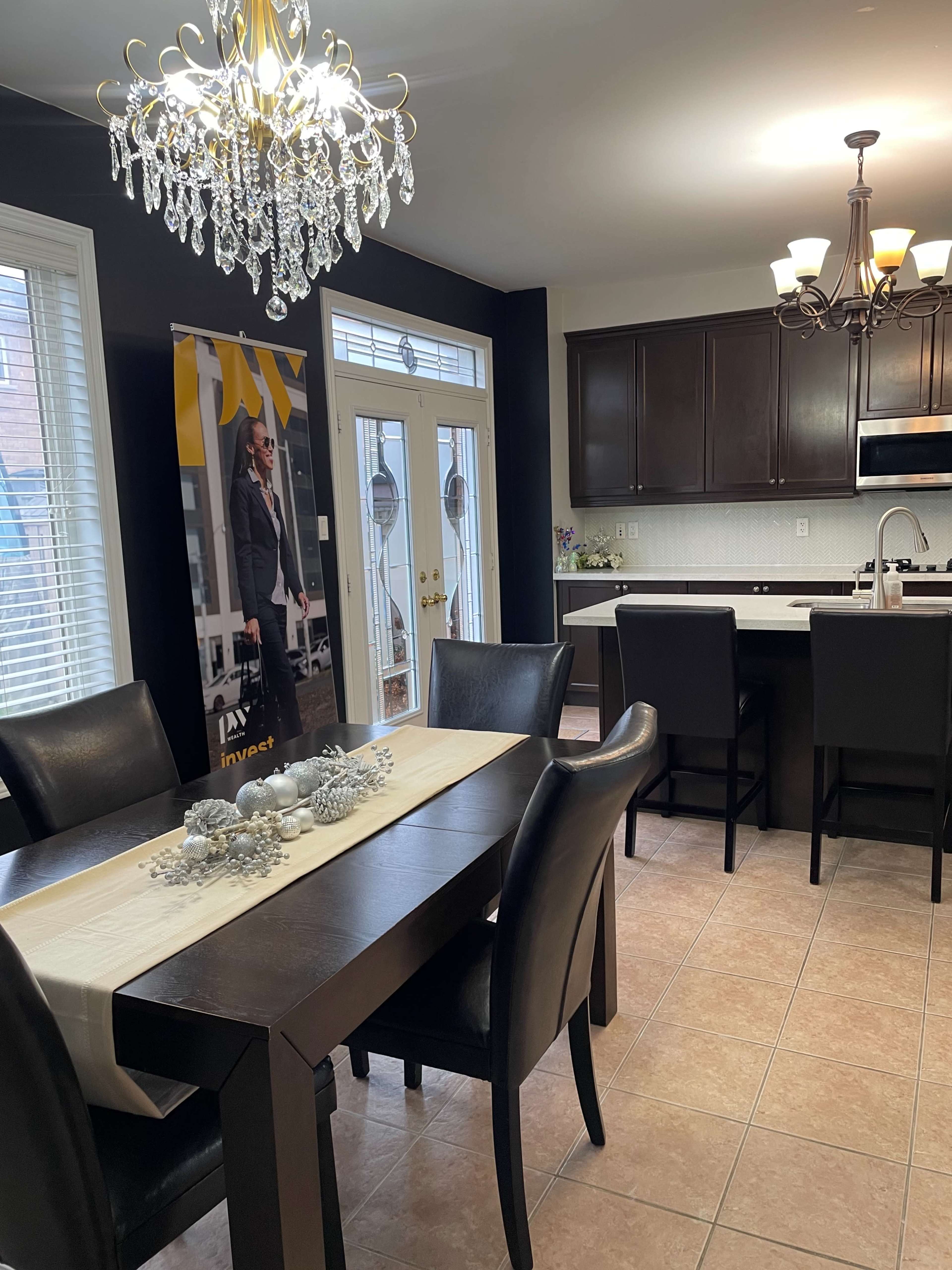 The image shows a modern kitchen and dining area with dark cabinetry, a chandelier, a table set for dining, and a large window letting in natural light.