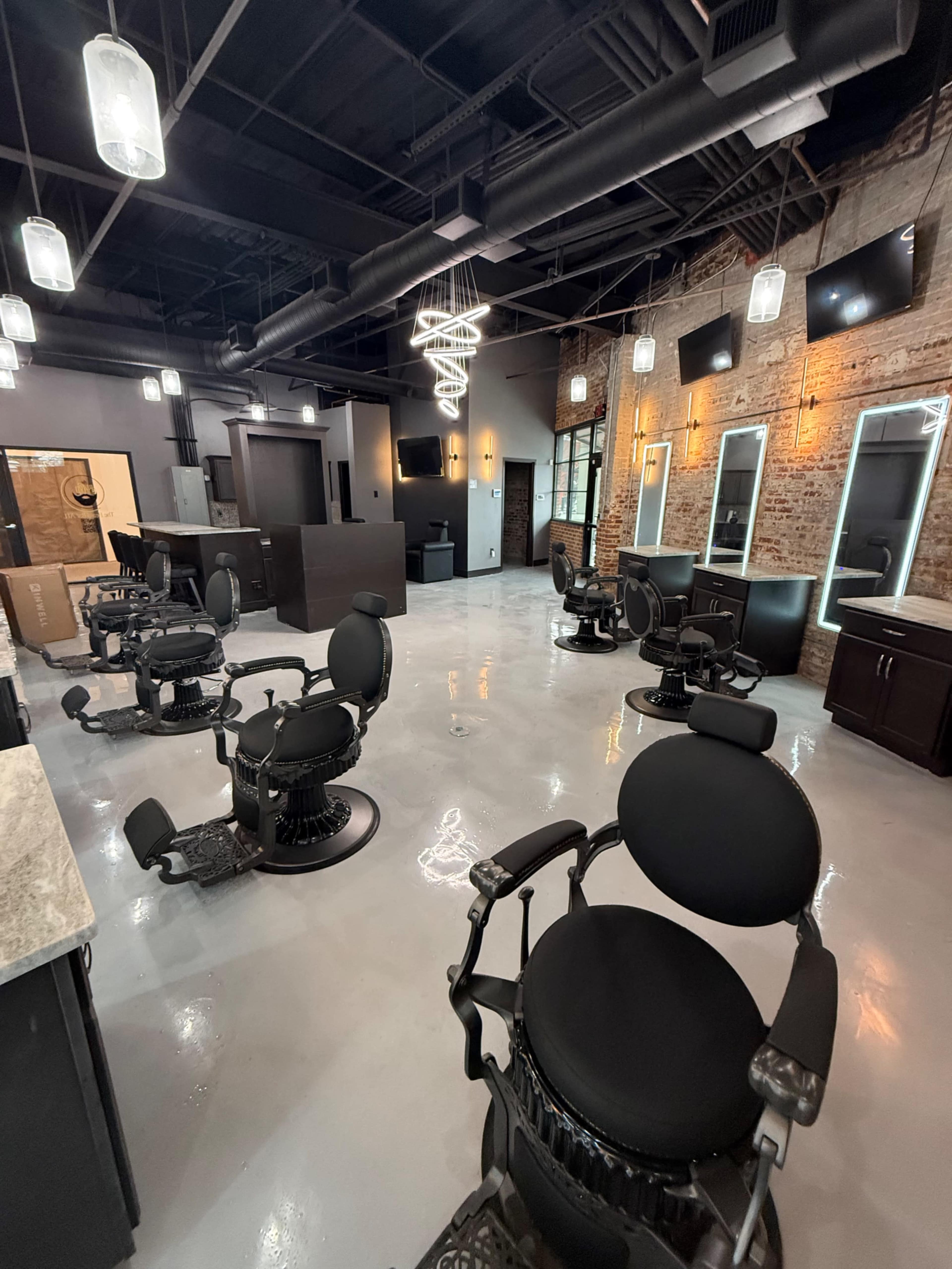 The image shows a modern hair salon with several barber chairs arranged in a spacious, well-lit interior featuring exposed brick walls and contemporary lighting fixtures.