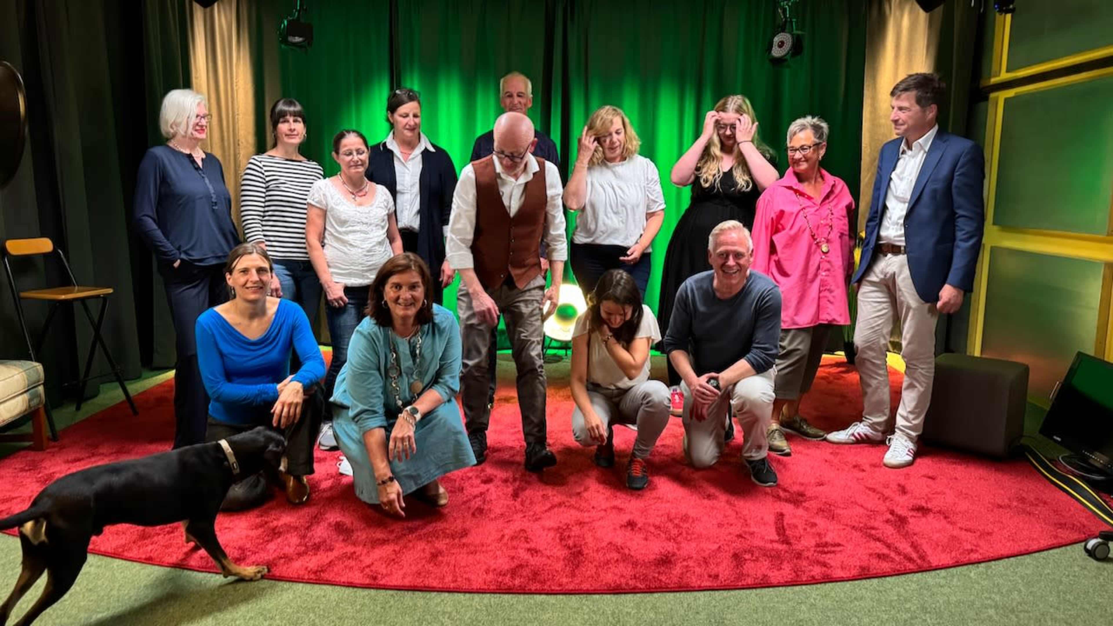 A diverse group of people, including men and women, poses together in a studio setting with green and yellow backdrop lighting, some smiling while others engage with cameras.