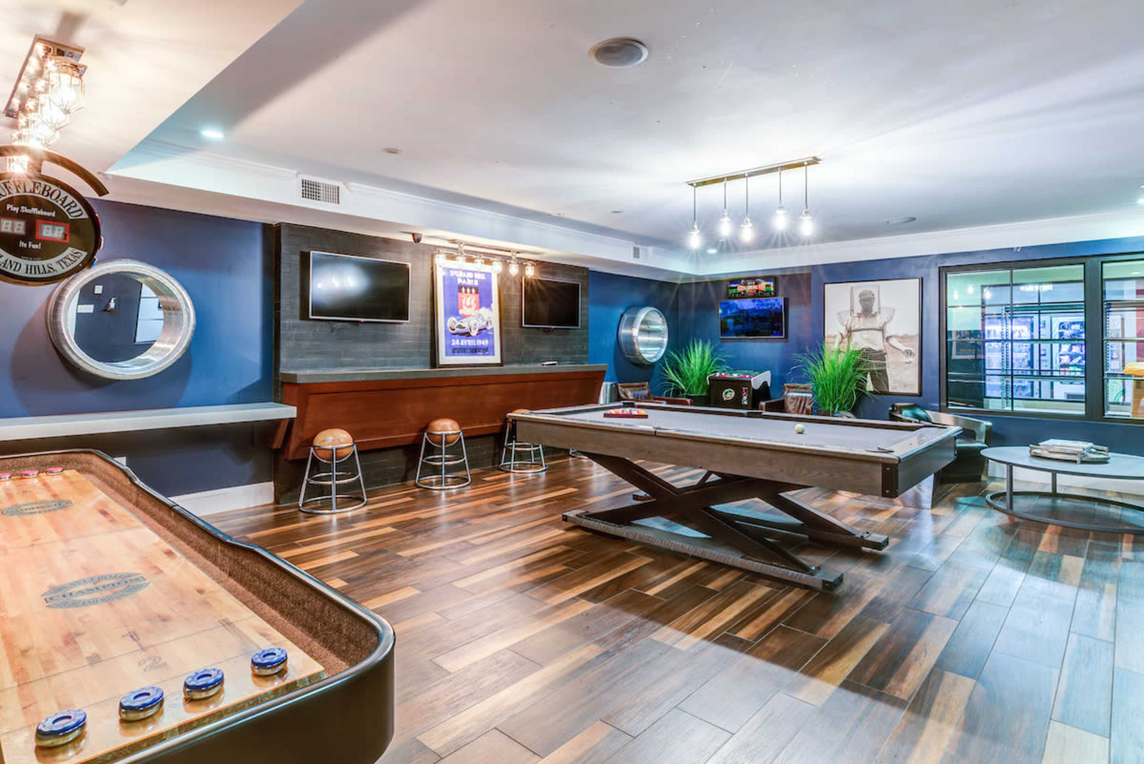 A recreational room featuring a pool table, a shuffleboard table, and several flat-screen TVs on the walls, with wooden flooring and modern lighting fixtures.