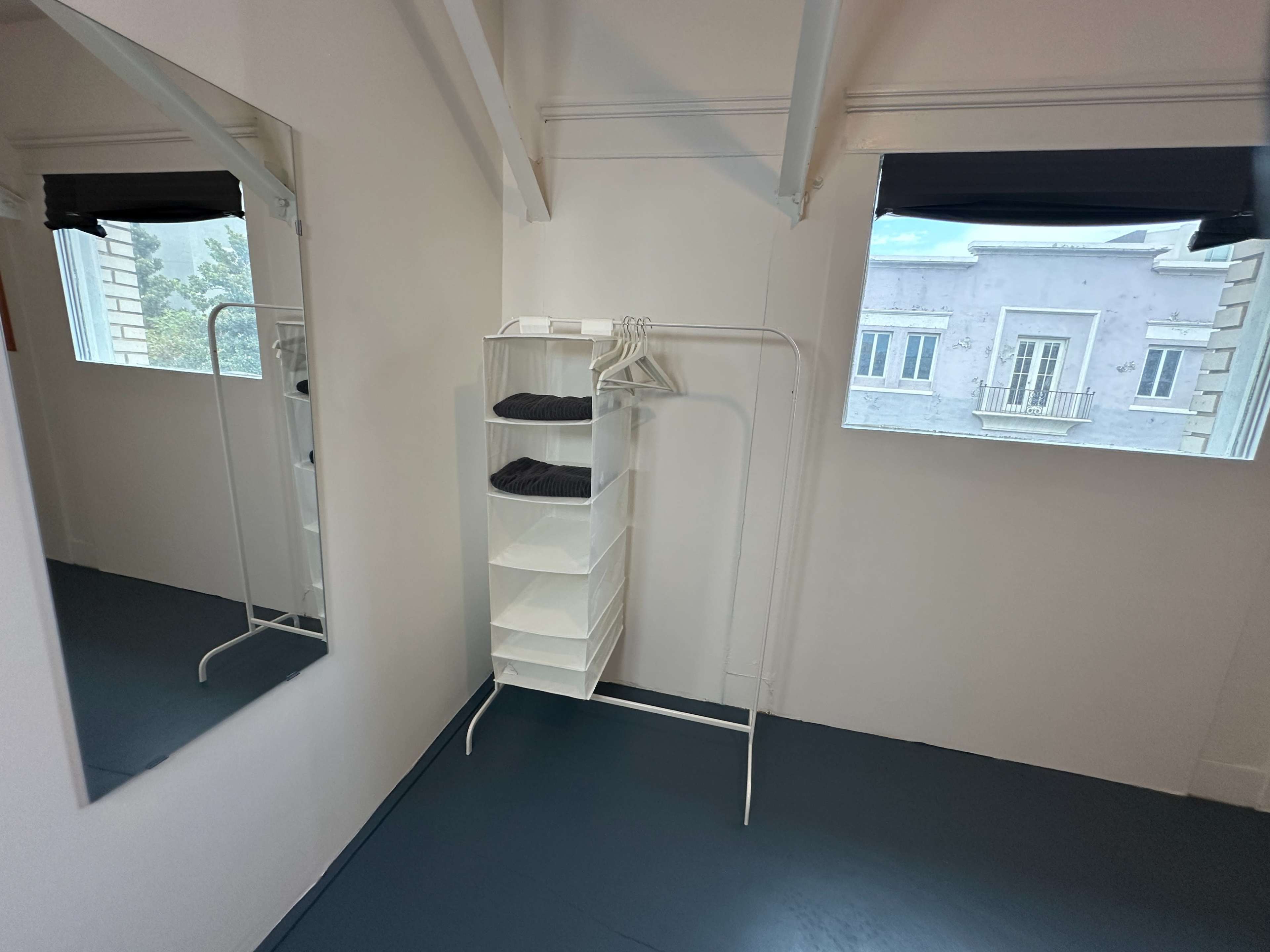 The image features a minimalist room with a white clothing rack and shelves, a large mirror, and a window showing a building exterior.