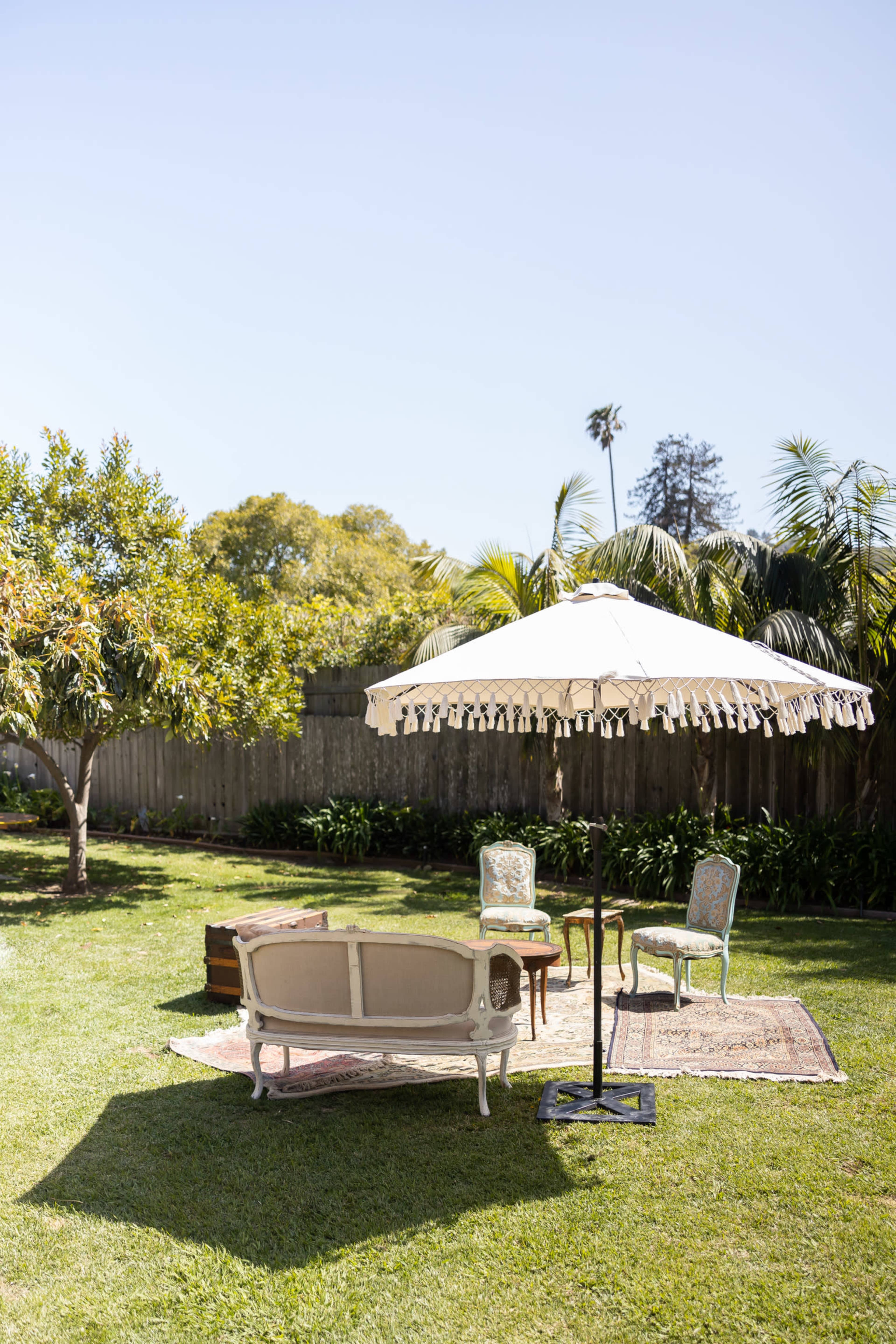 A sunlit backyard features a vintage couch, a small table, and a large umbrella on a patterned rug surrounded by greenery.