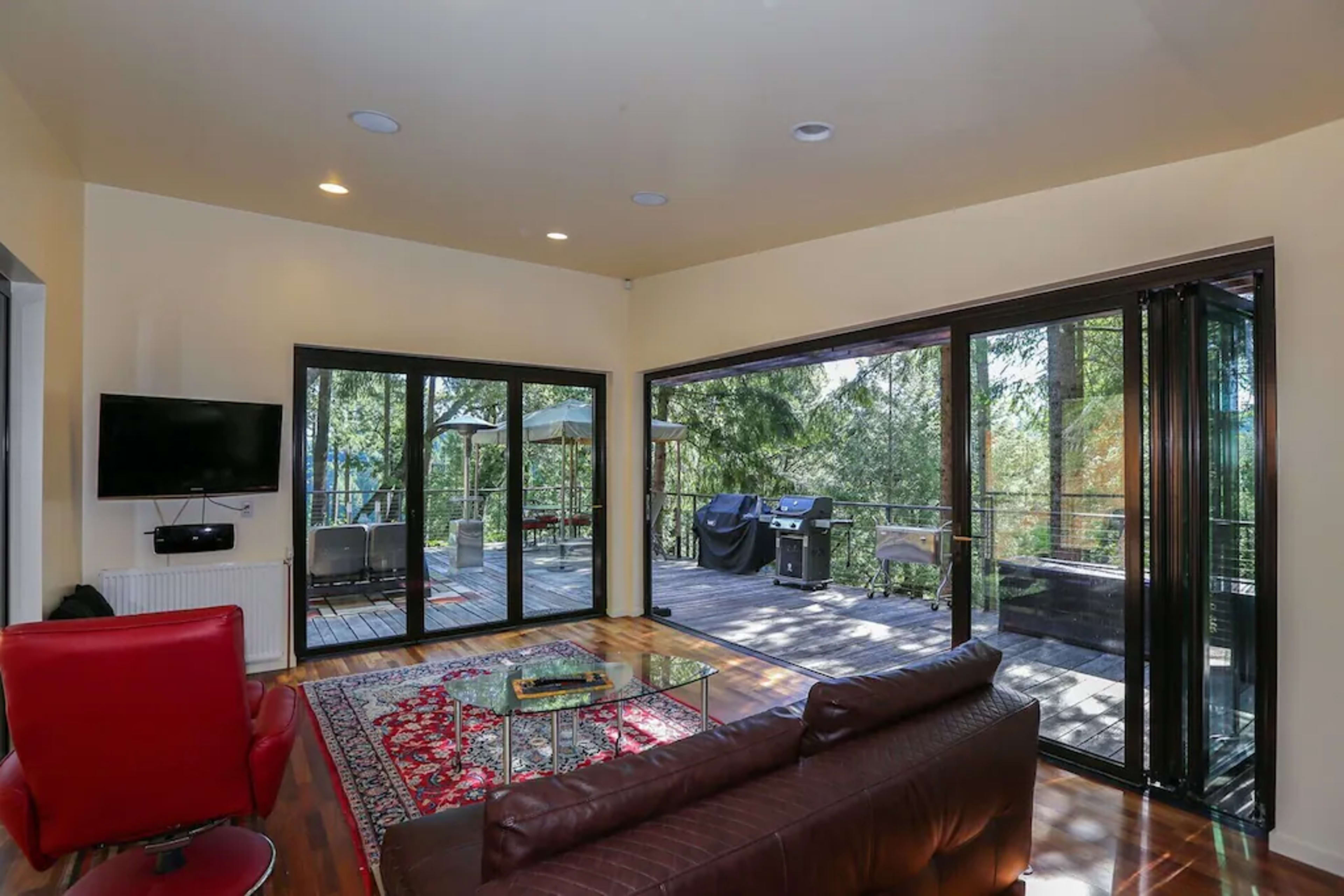 A modern living room features large glass doors opening to a wooden deck surrounded by trees, with a red chair and a brown sofa inside.