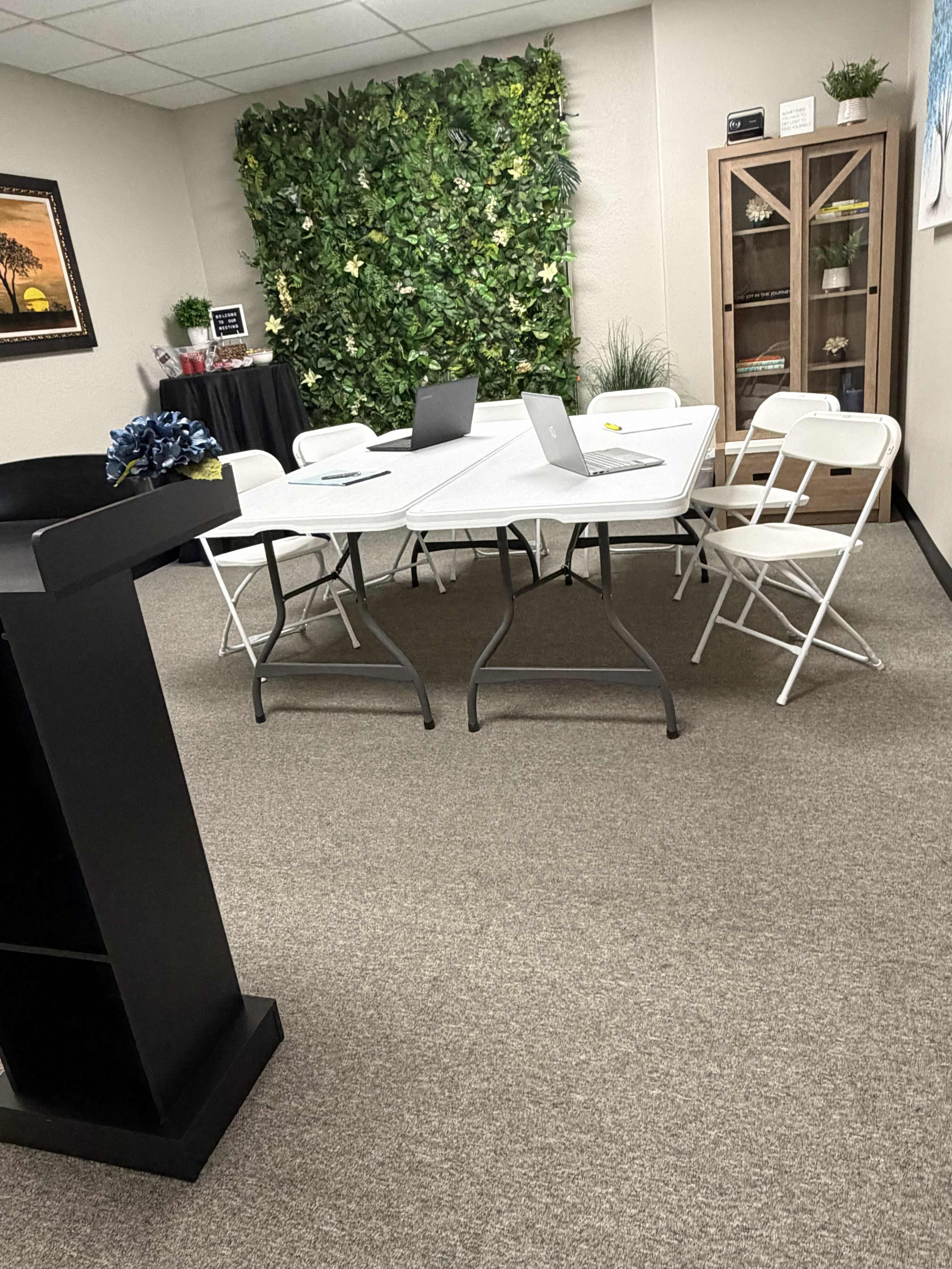 A small meeting room features a table with laptops and several white folding chairs, set against a wall with artificial greenery and a cabinet.