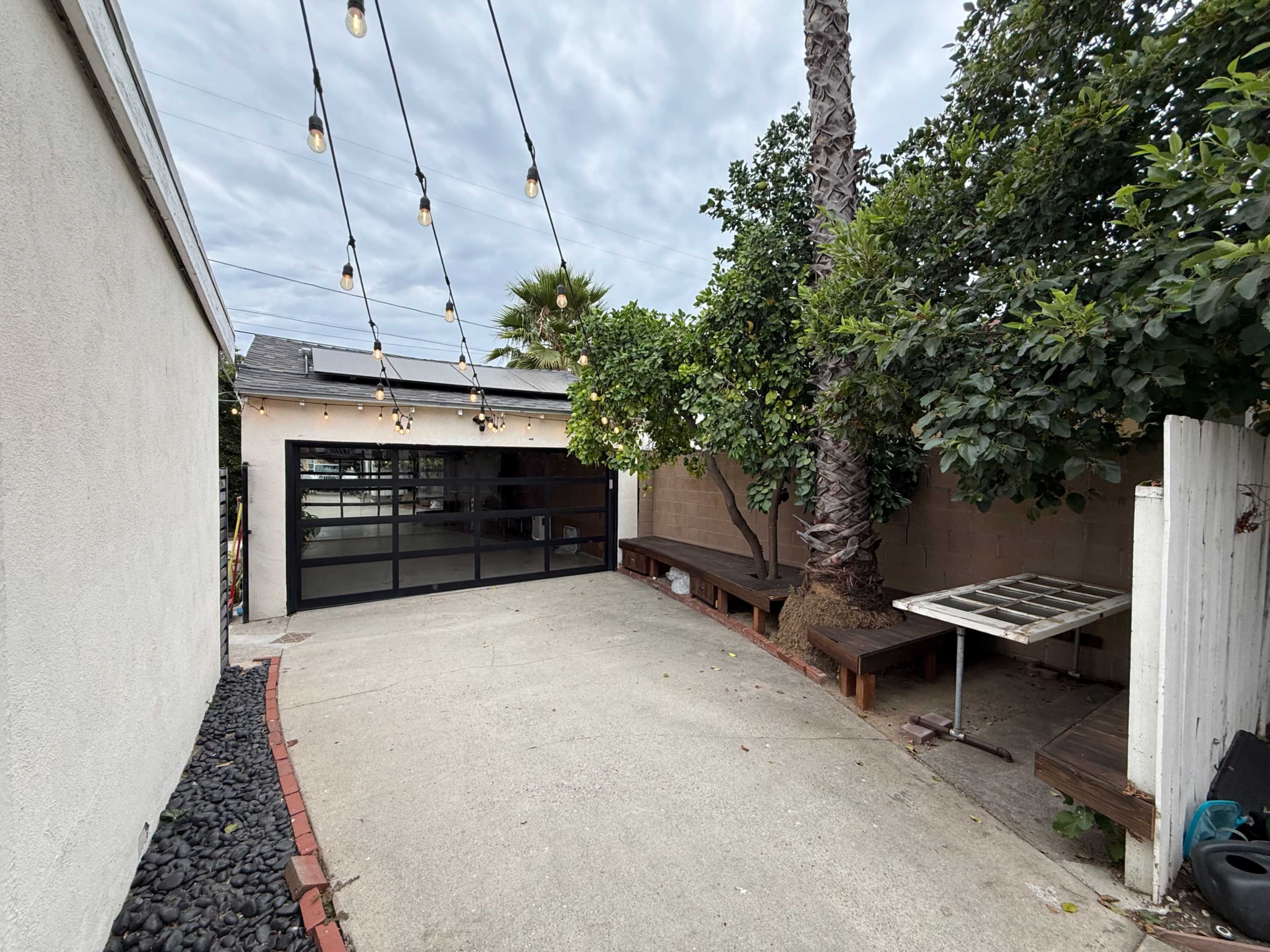 The image shows a concrete driveway leading to a garage door, surrounded by trees and string lights above.