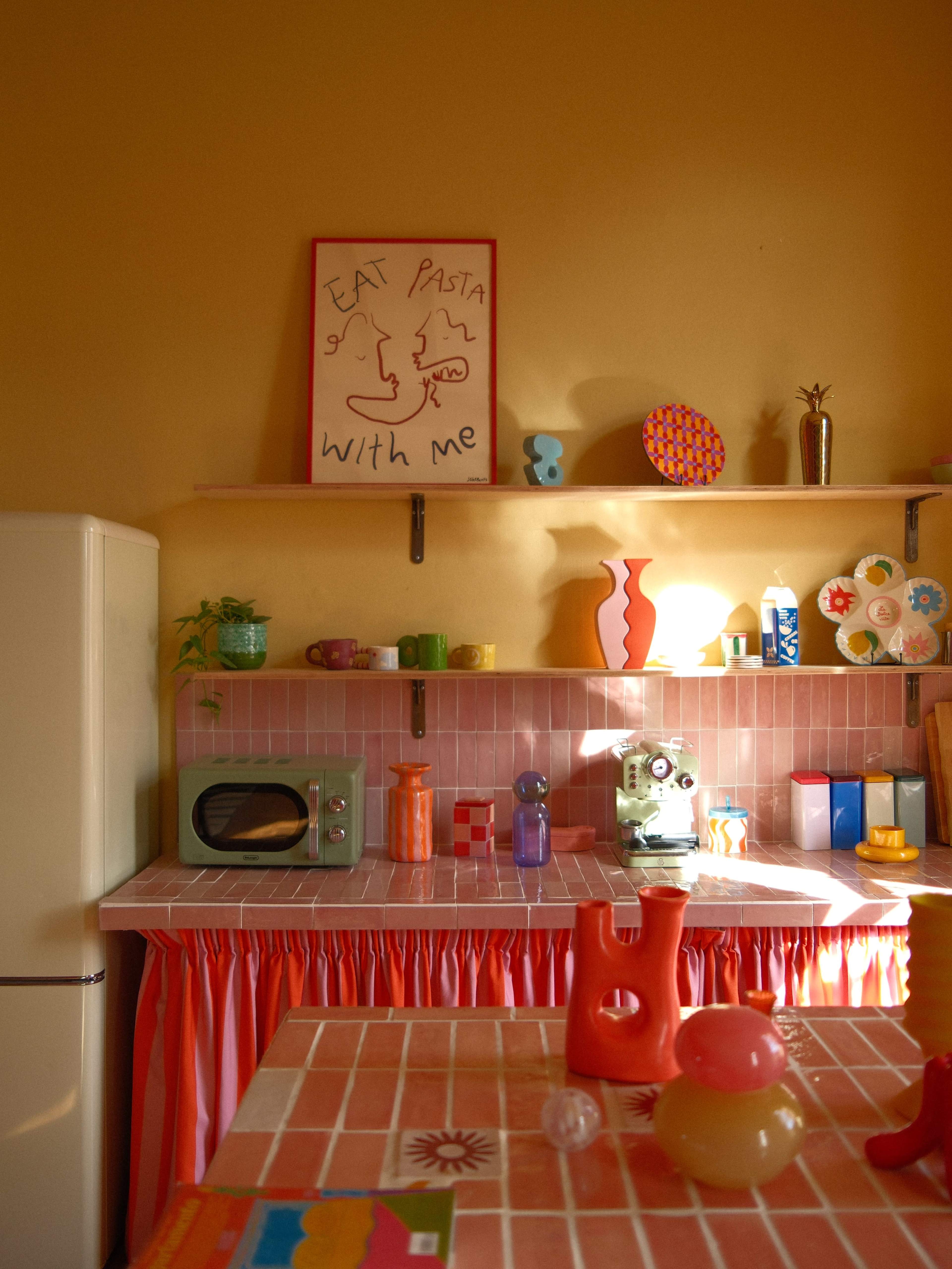 The image depicts a colorful kitchen with a pink tiled countertop, various kitchen utensils and decorations, and a framed sign that reads "EAT PASTA WITH ME" hanging above.