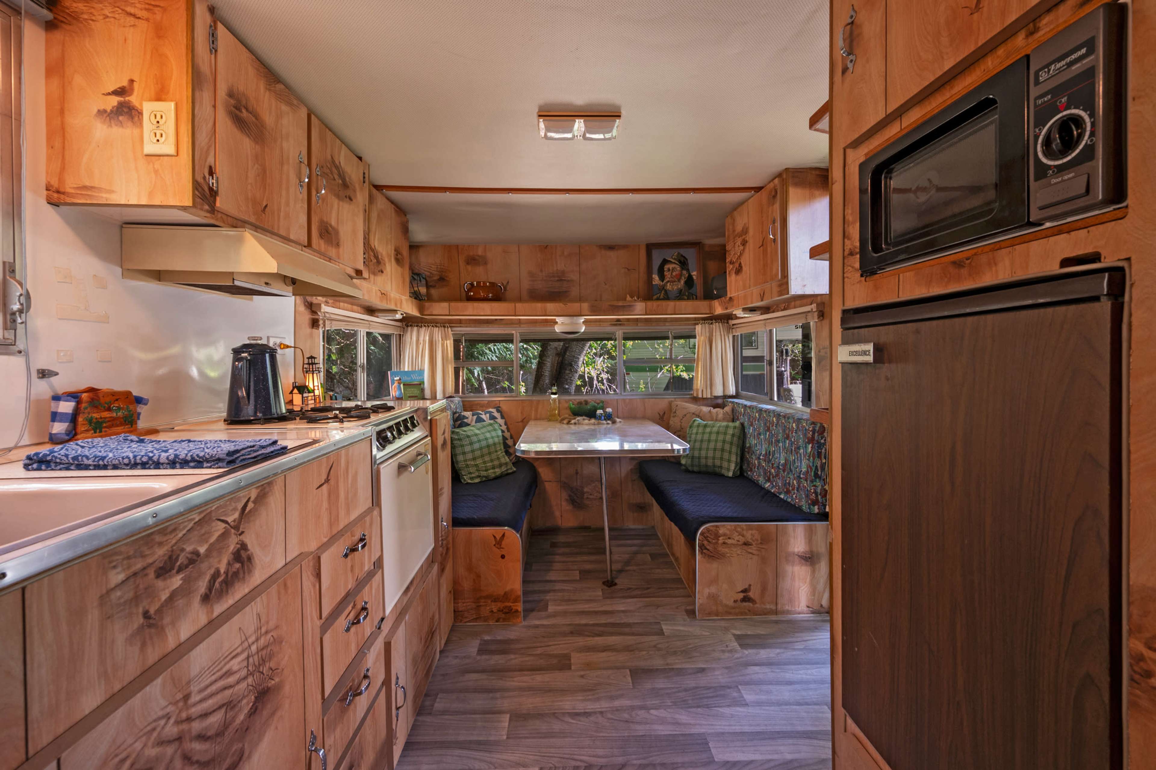 The interior of a vintage camper features wooden cabinets, a kitchenette with a stove, and a dining area with cushioned benches.