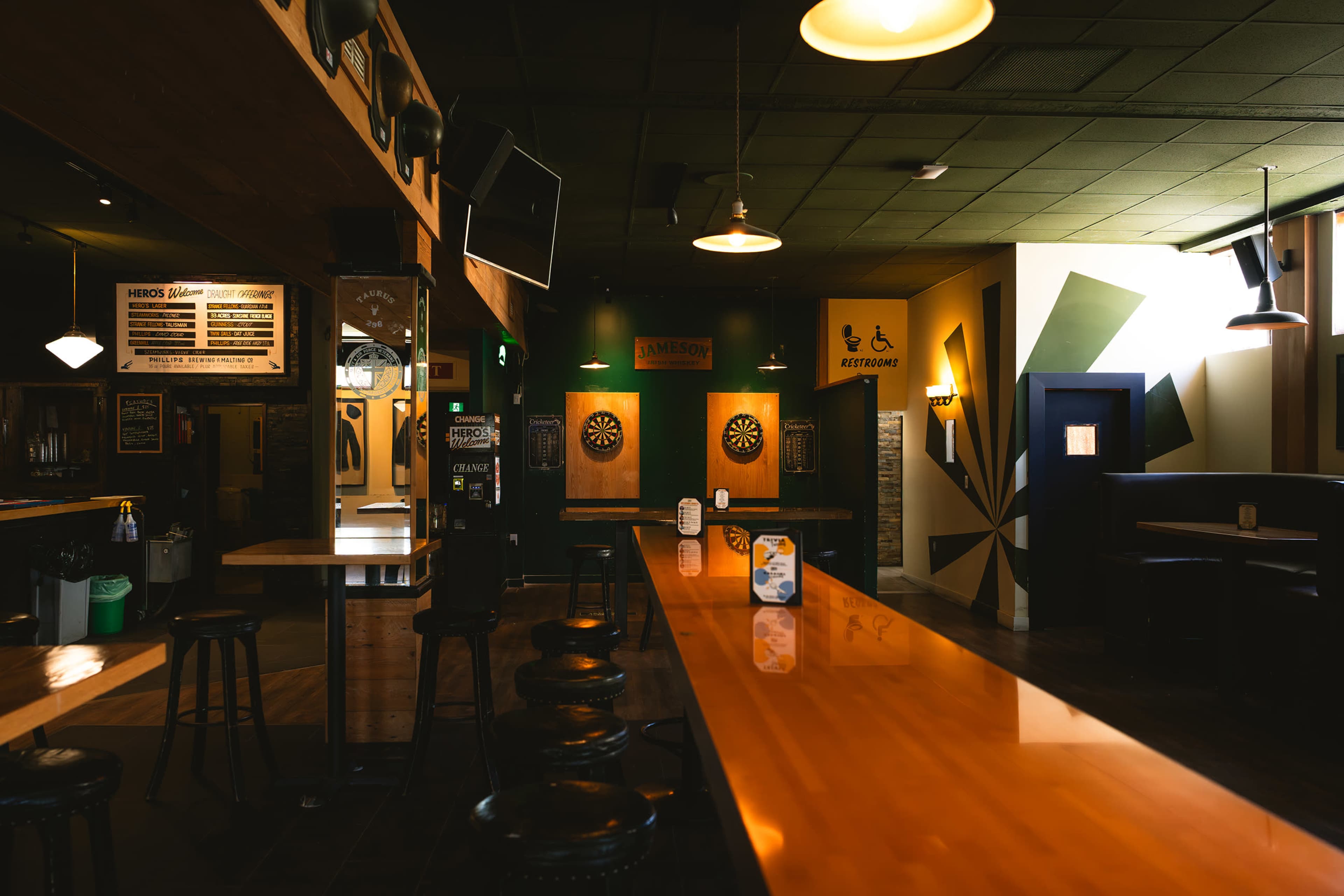 The image shows a spacious bar area featuring a long wooden counter, dartboards on the walls, and a few tables arranged throughout the room.