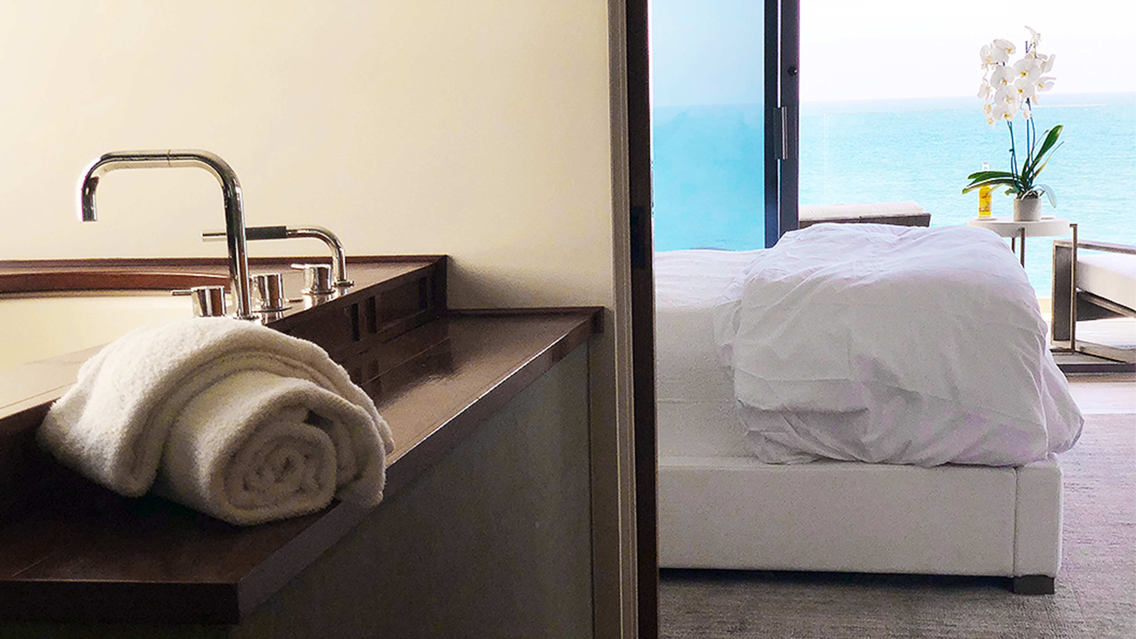 A bathroom area with rolled towels on a countertop, leading to a spacious room with a bed and a view of the ocean through an open door.