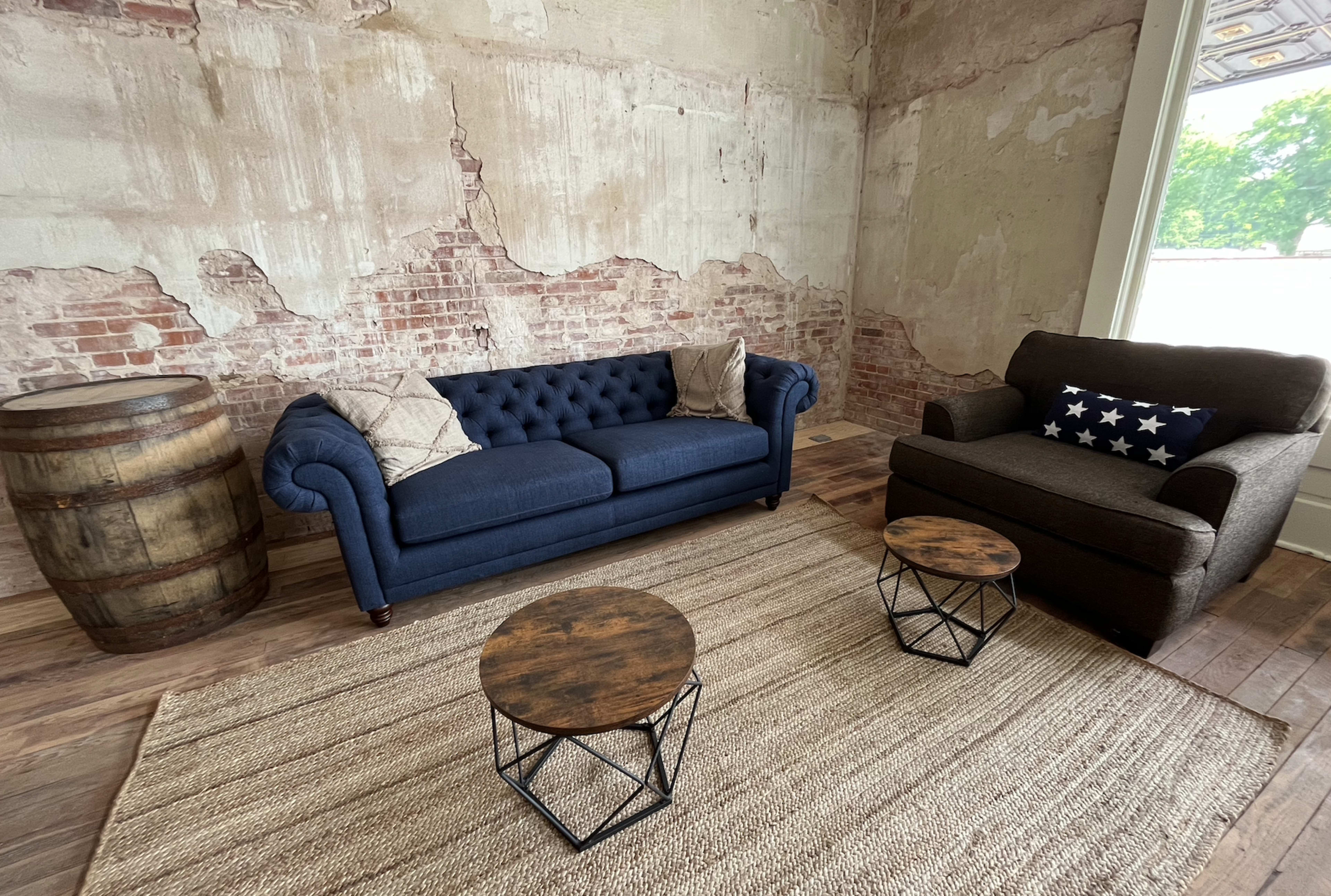 The image shows a sitting area with a blue tufted sofa, a brown armchair, two small wooden tables, and a barrel beside them on a woven rug, against a backdrop of exposed brick walls.