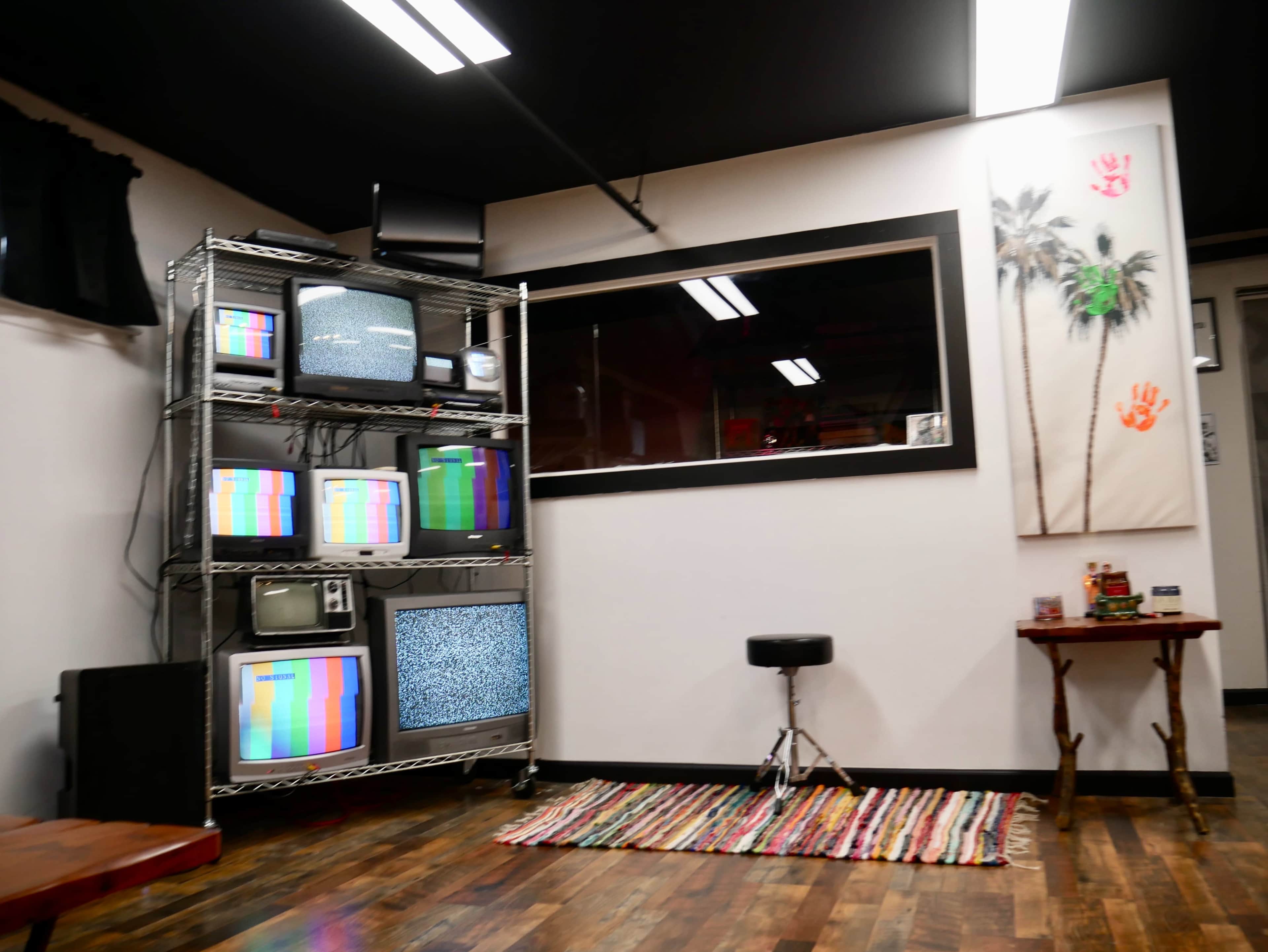 The image shows a room with multiple vintage televisions on a metal rack, a small stool, and a colorful woven rug on the wooden floor.