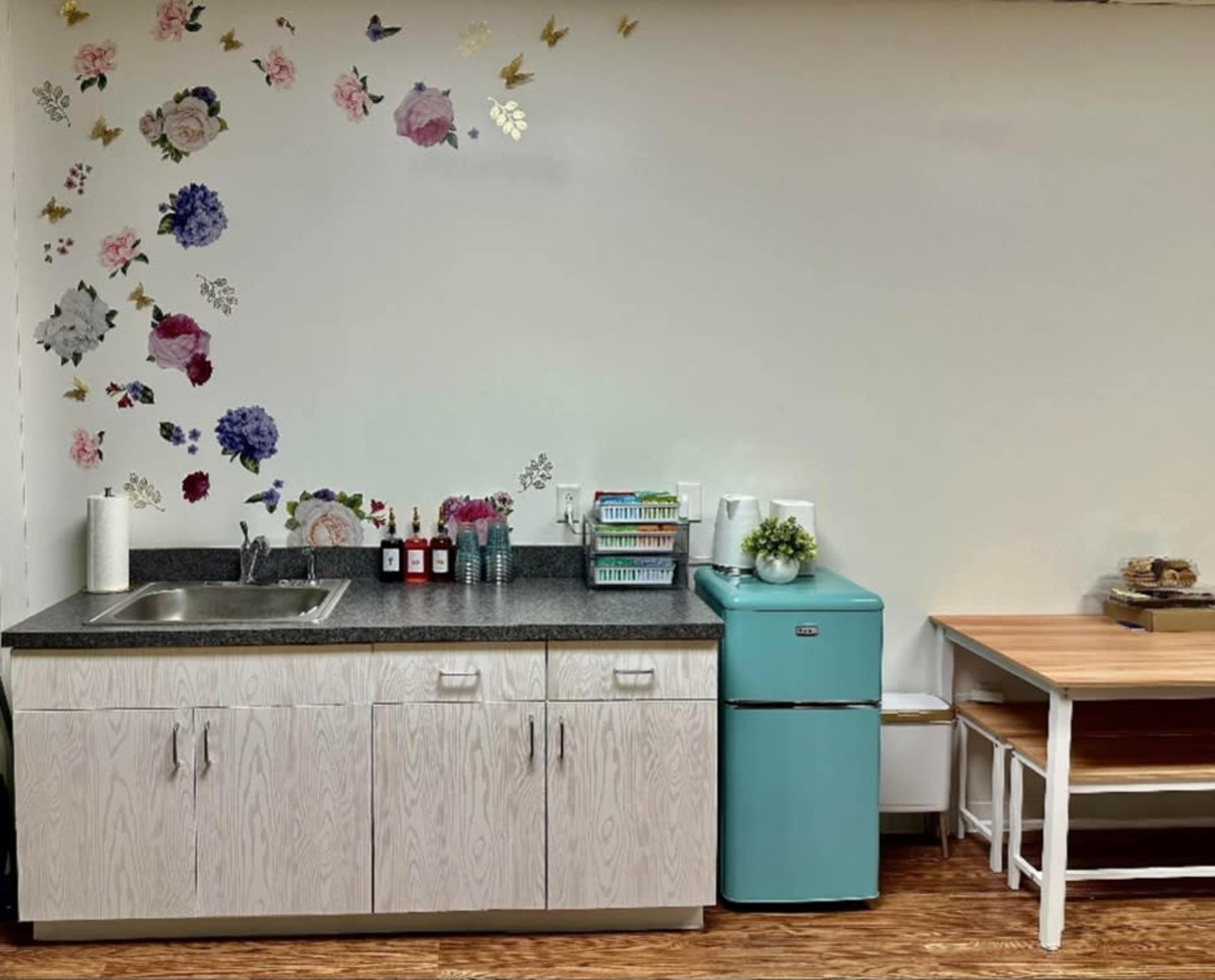 A small kitchen area features a sink, a turquoise fridge, and a wooden table, with floral decorations on the wall.