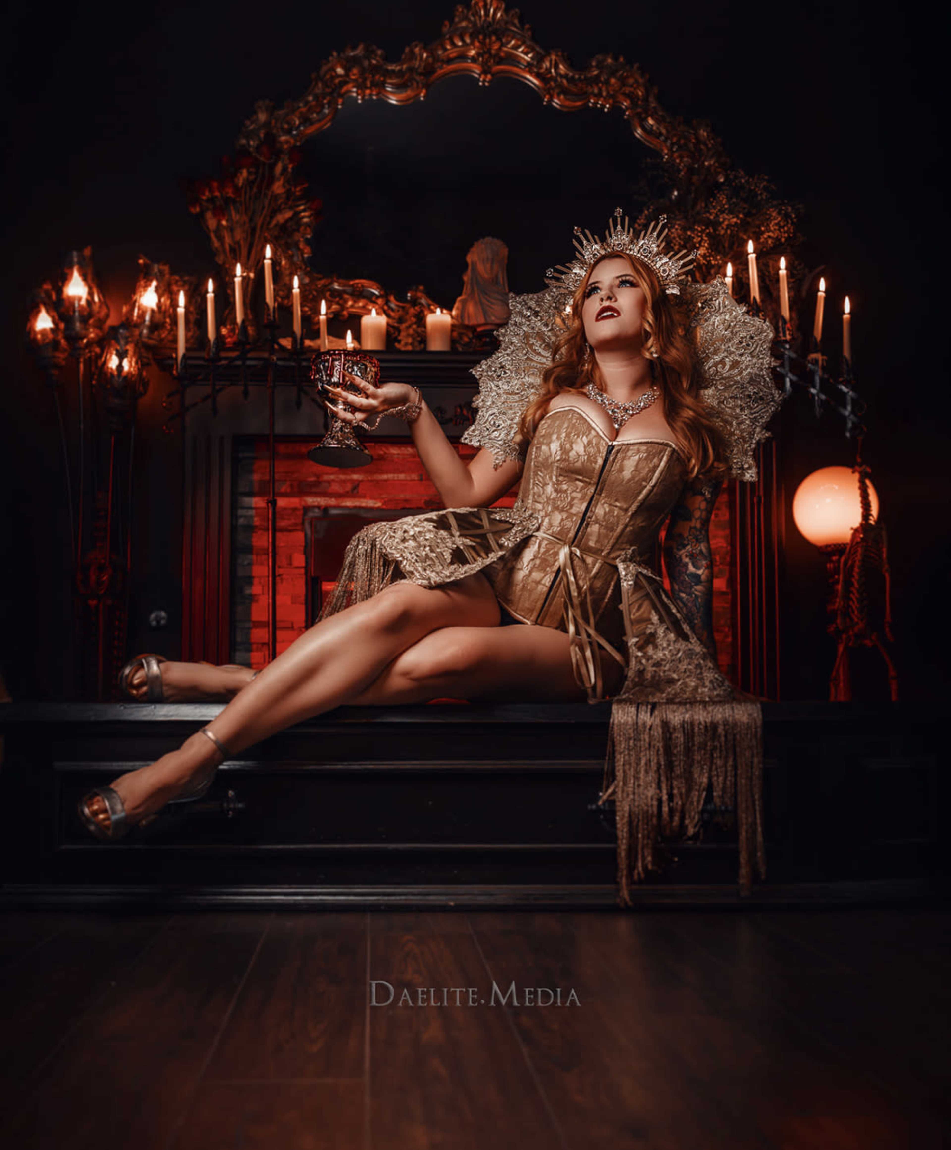 A woman in a gold corset sits on an ornate throne surrounded by candles, holding a goblet in a dark, elegantly decorated room.