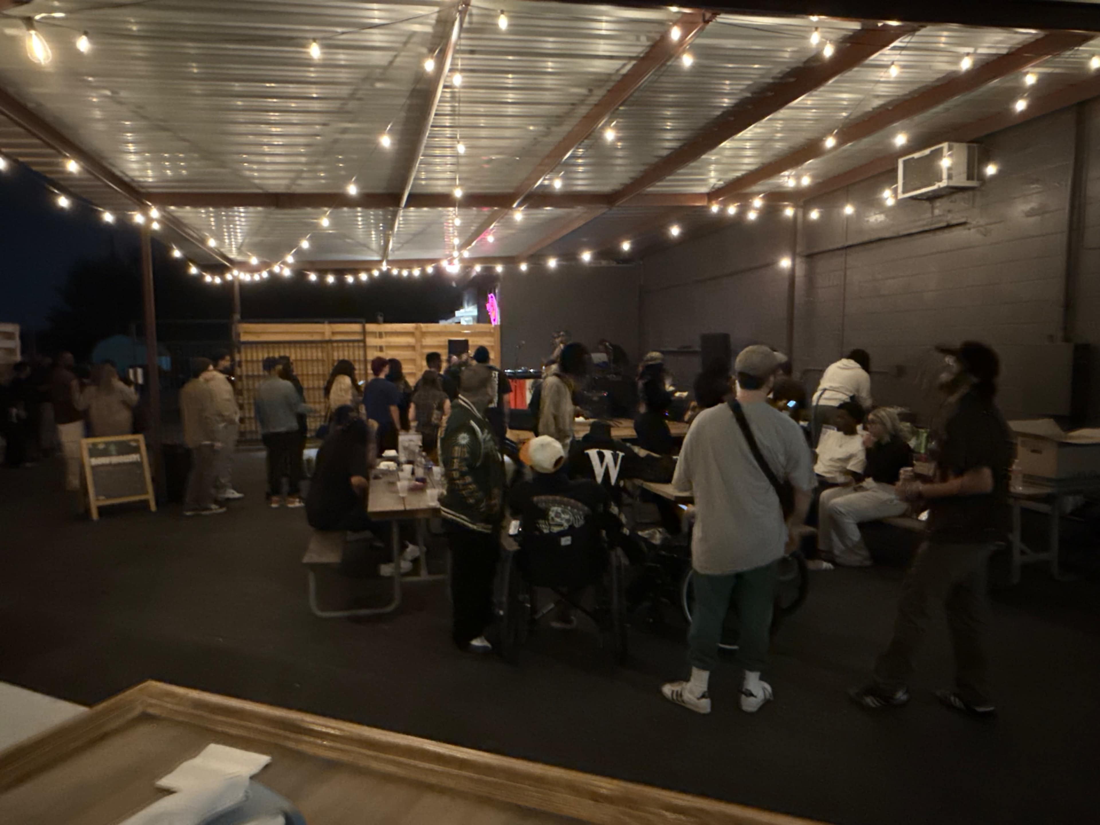 A crowd gathers under a covered outdoor area with string lights, enjoying music and conversation at an event.