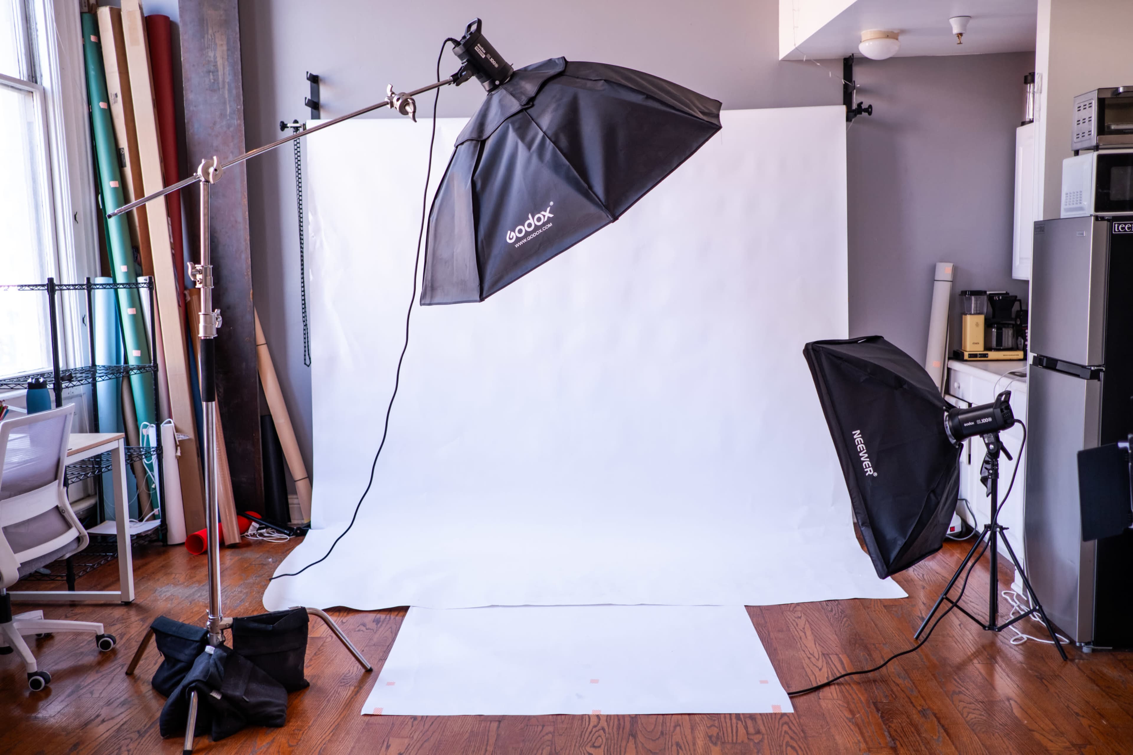 A photography studio is set up with softbox lights, a white backdrop, and various equipment including a chair and kitchen appliances in the background.