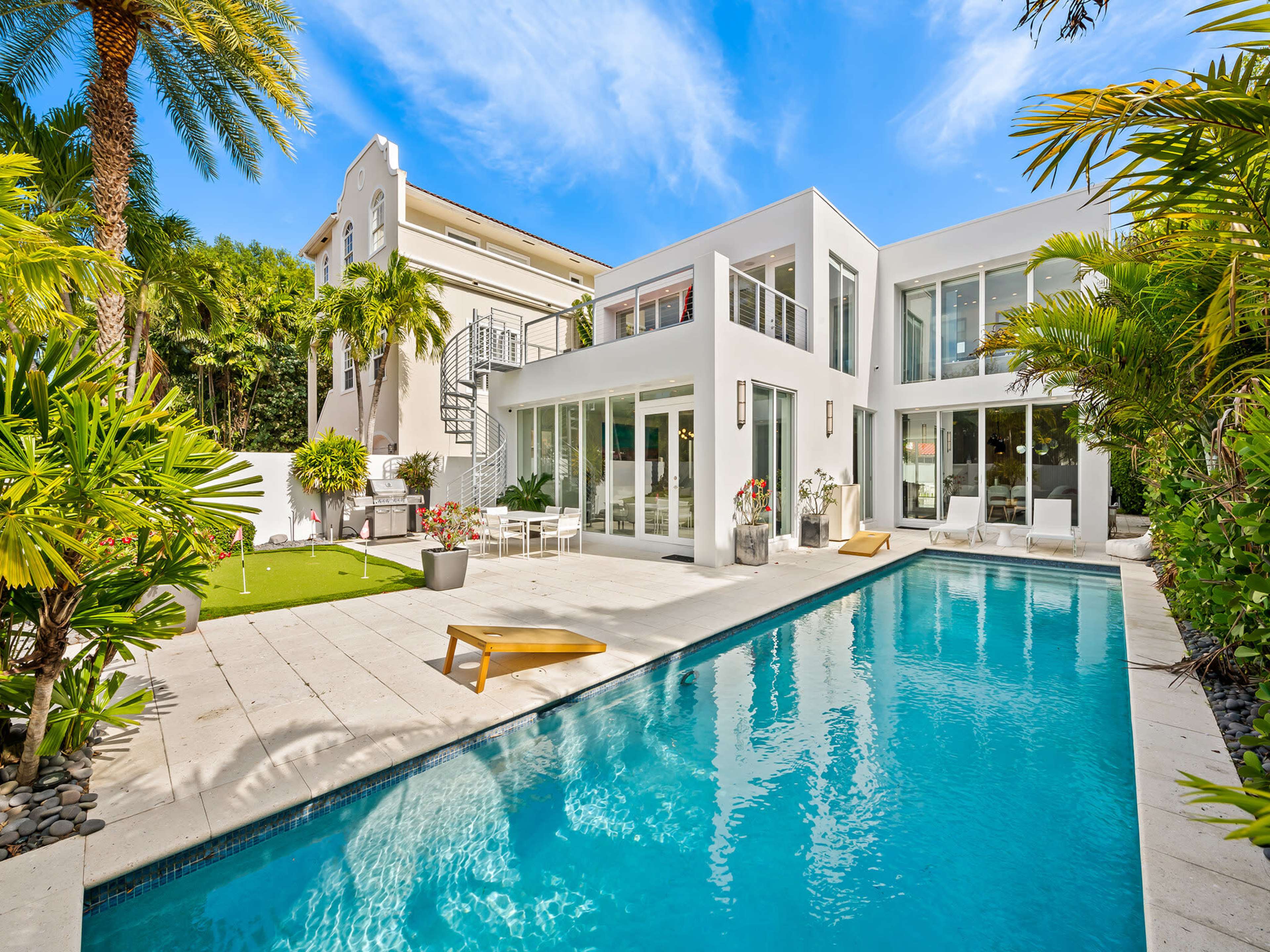 The image showcases a modern white house with large windows, a swimming pool, and palm trees in the backyard.