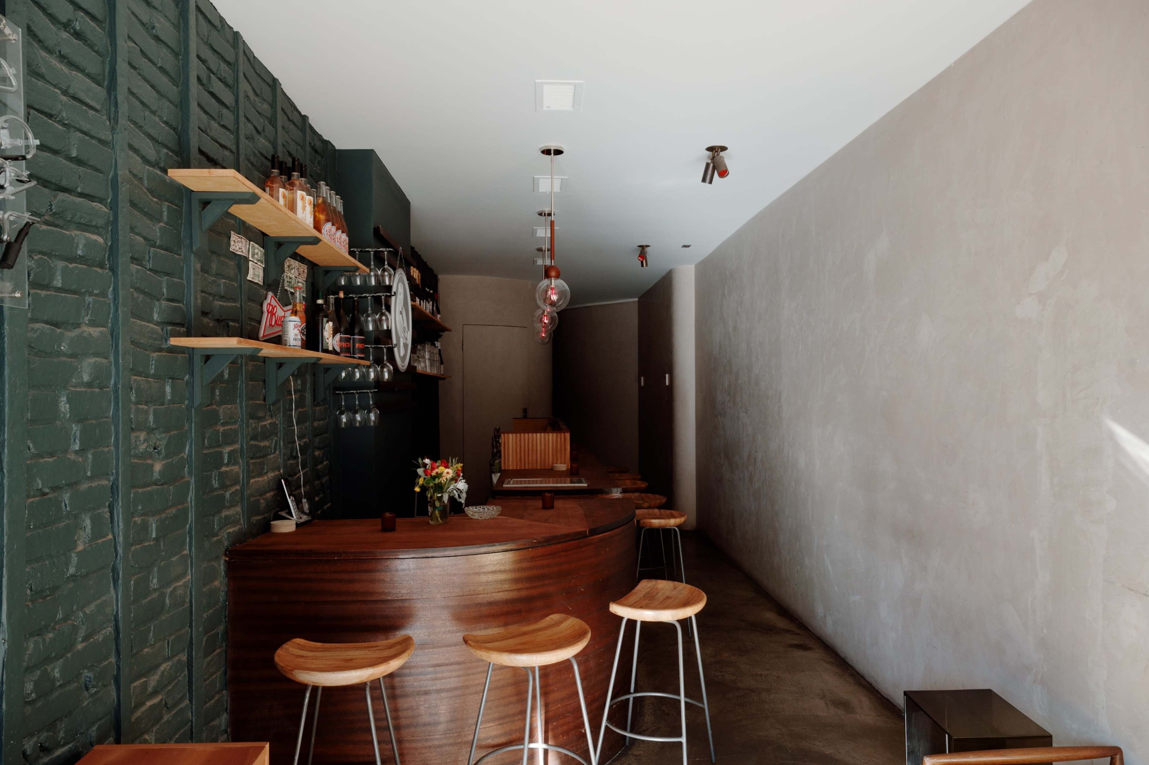 The image shows a minimalist bar with wooden stools, a curved counter, and shelves displaying various bottles along a green brick wall.