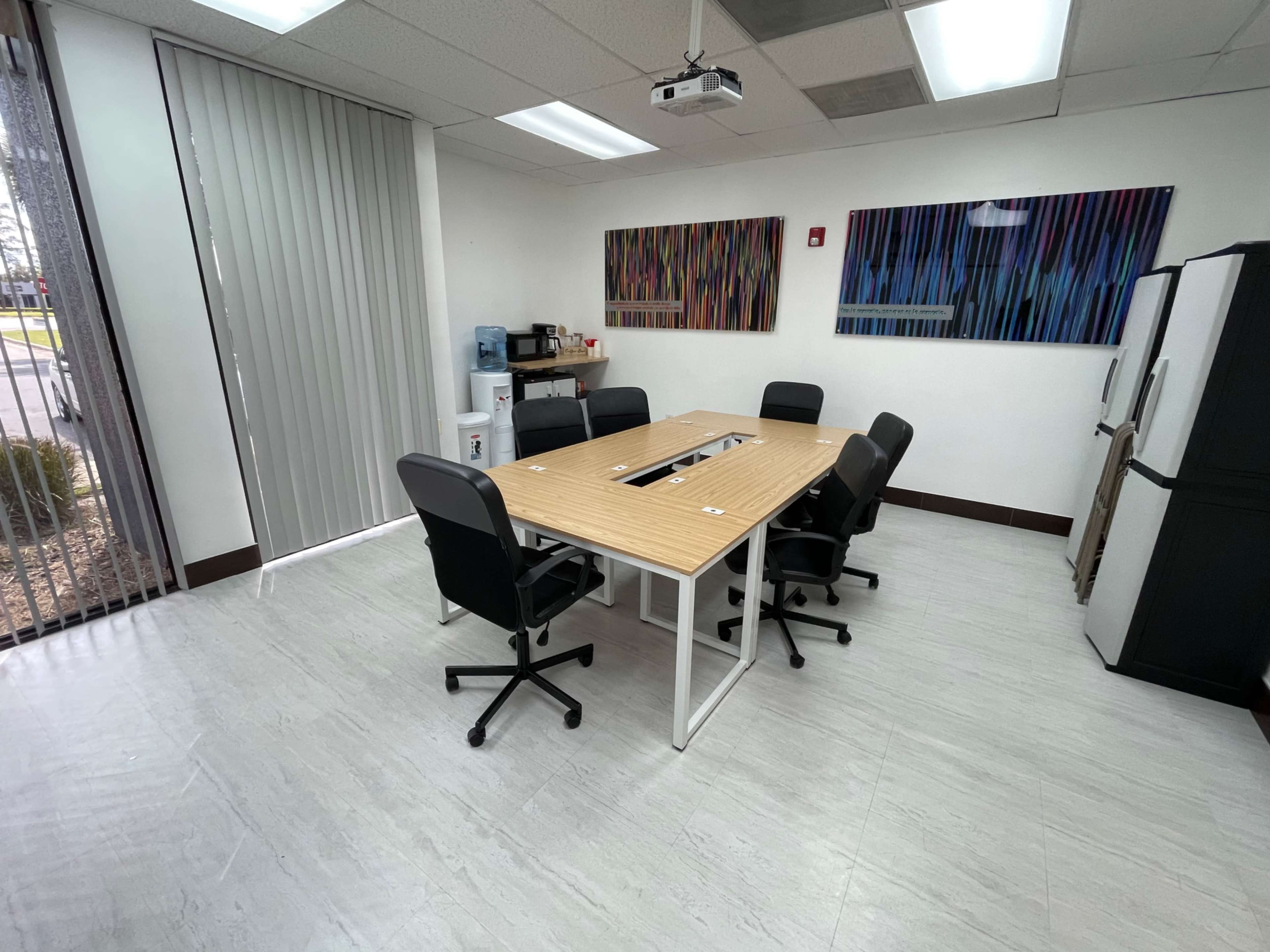 A modern conference room with a wooden table, six black chairs, a refrigerator, and colorful artwork on the walls.