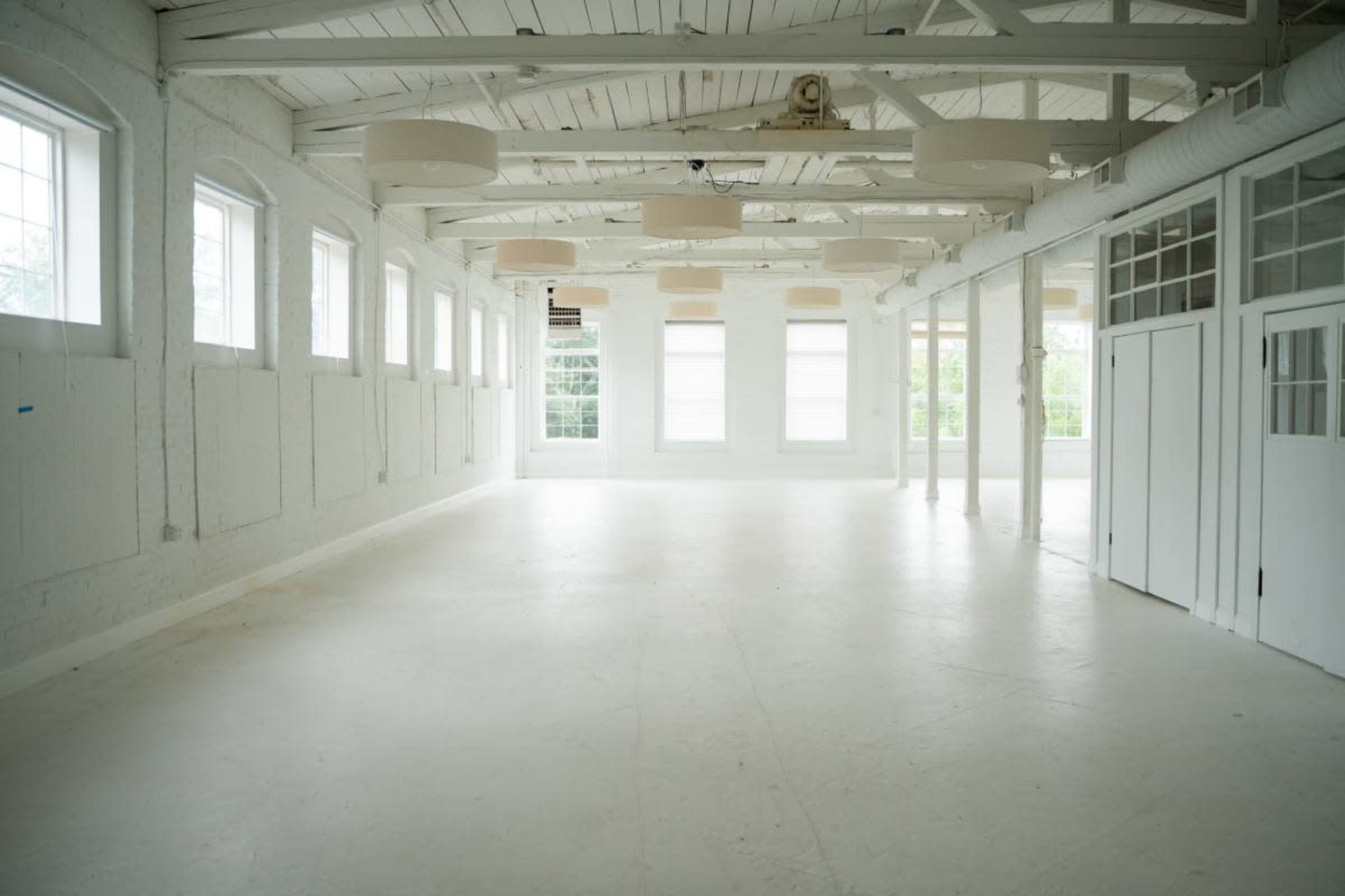 The image shows a large, empty room with white walls, a wooden ceiling, and multiple windows allowing natural light to enter.