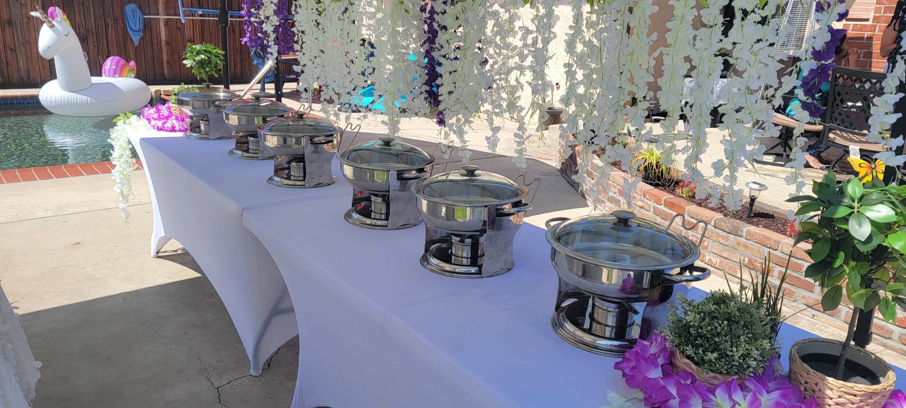 A long table is set up by a pool, featuring several silver chafing dishes and floral decorations hanging above.