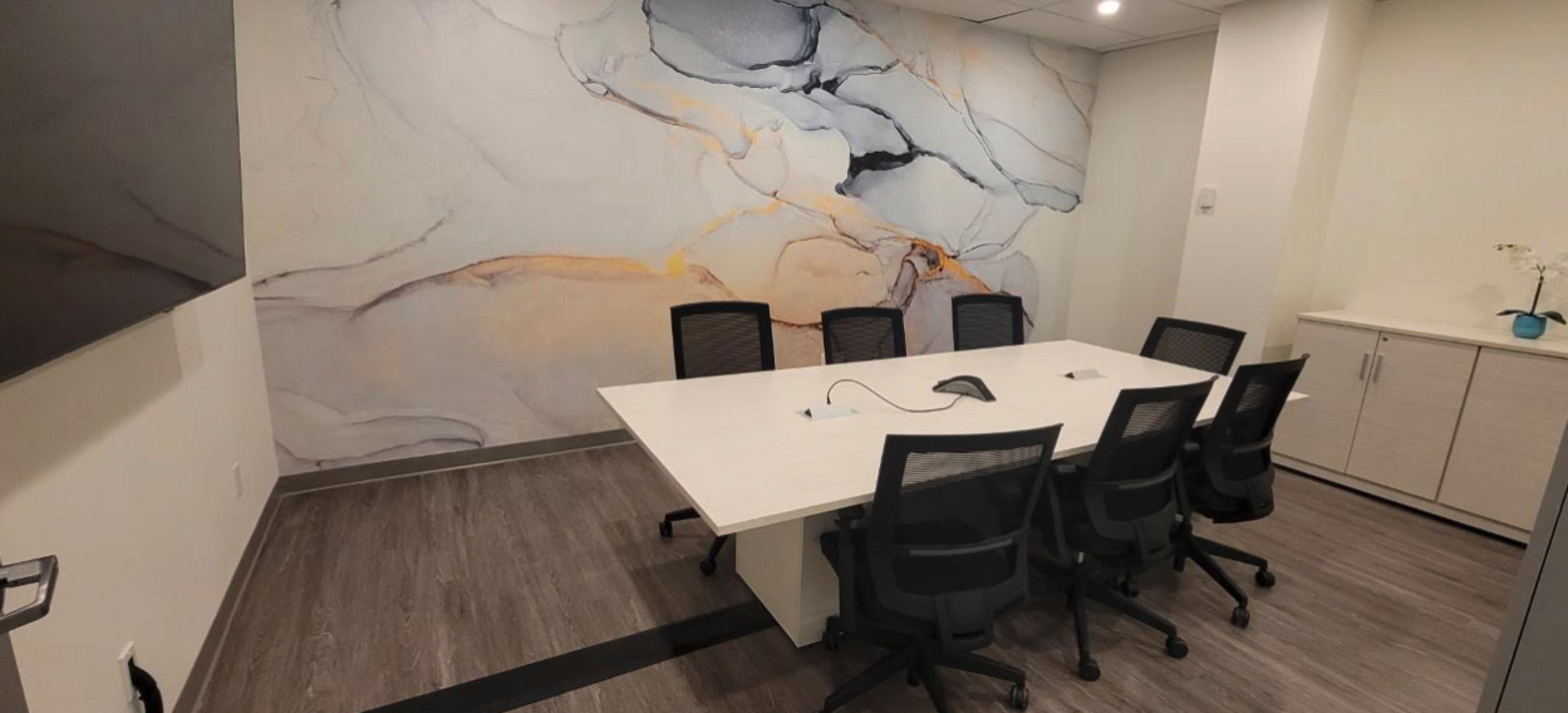 A modern conference room features a large white table surrounded by black chairs, with a mural of abstract shapes on the wall behind it.