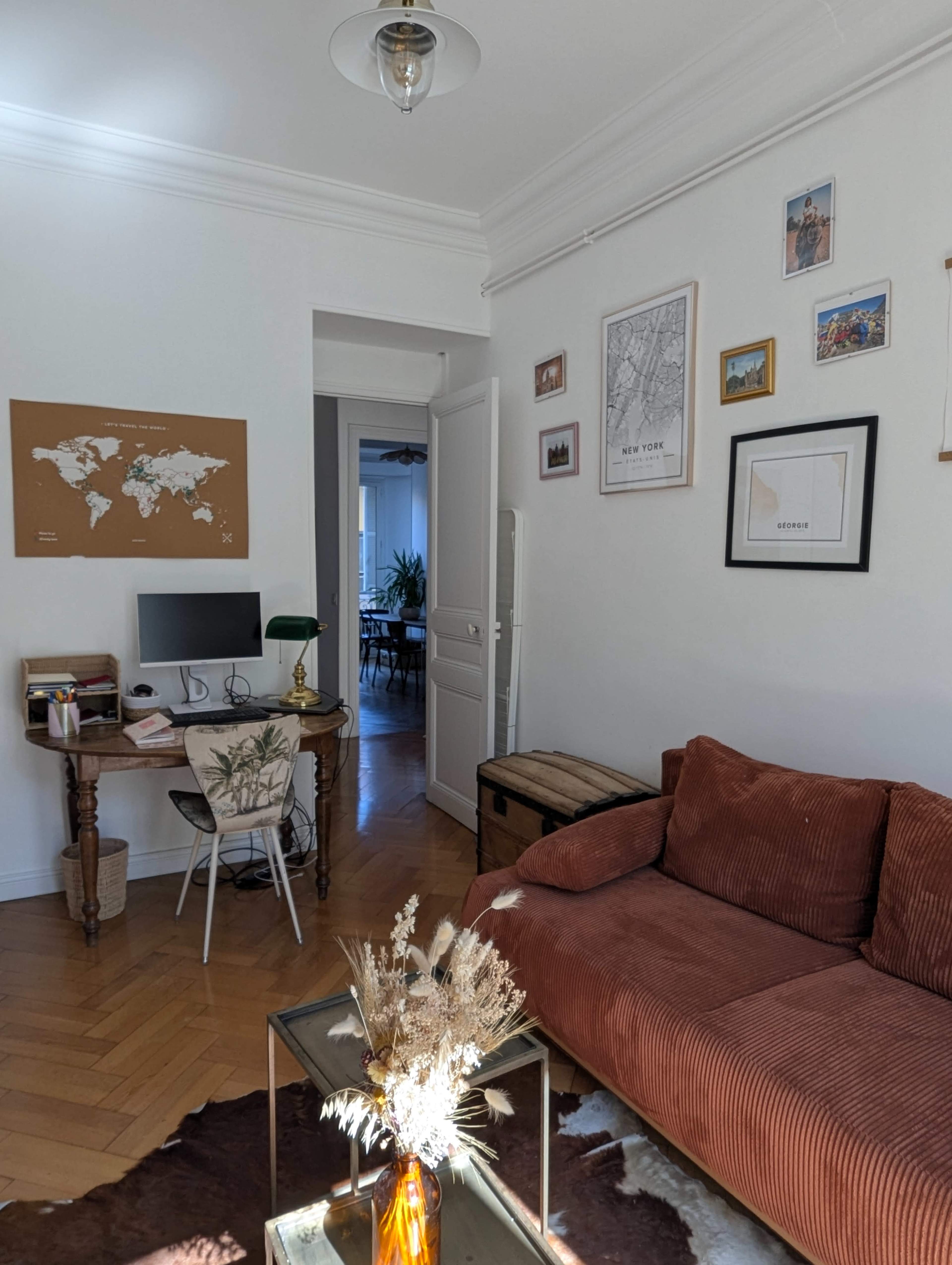 The image shows a cozy living room with a brown sofa, a desk with a computer, and various framed pictures on the walls.