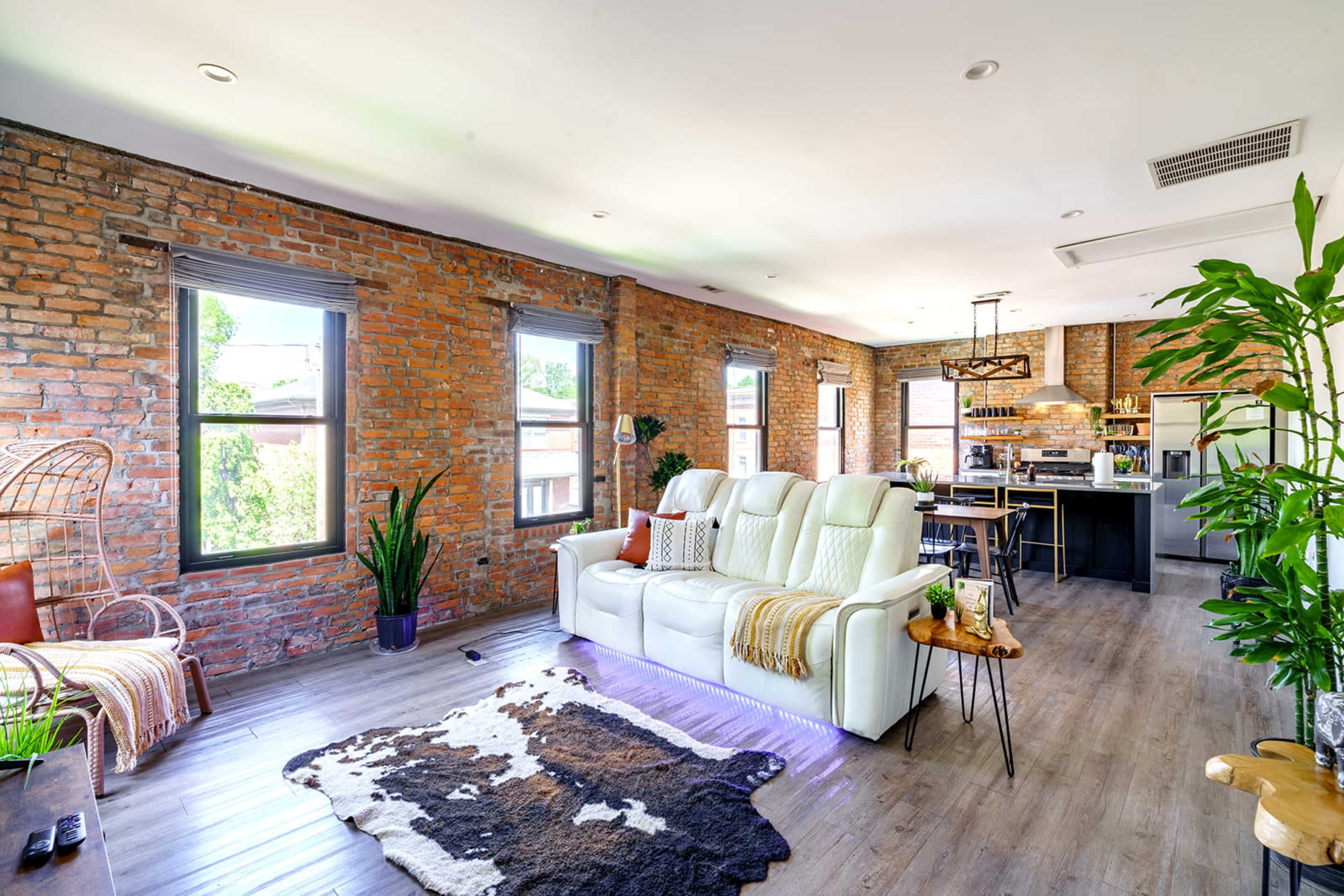 The image shows a modern living room with exposed brick walls, a white sectional sofa, and a view into a kitchen area featuring an island and pendant lighting.