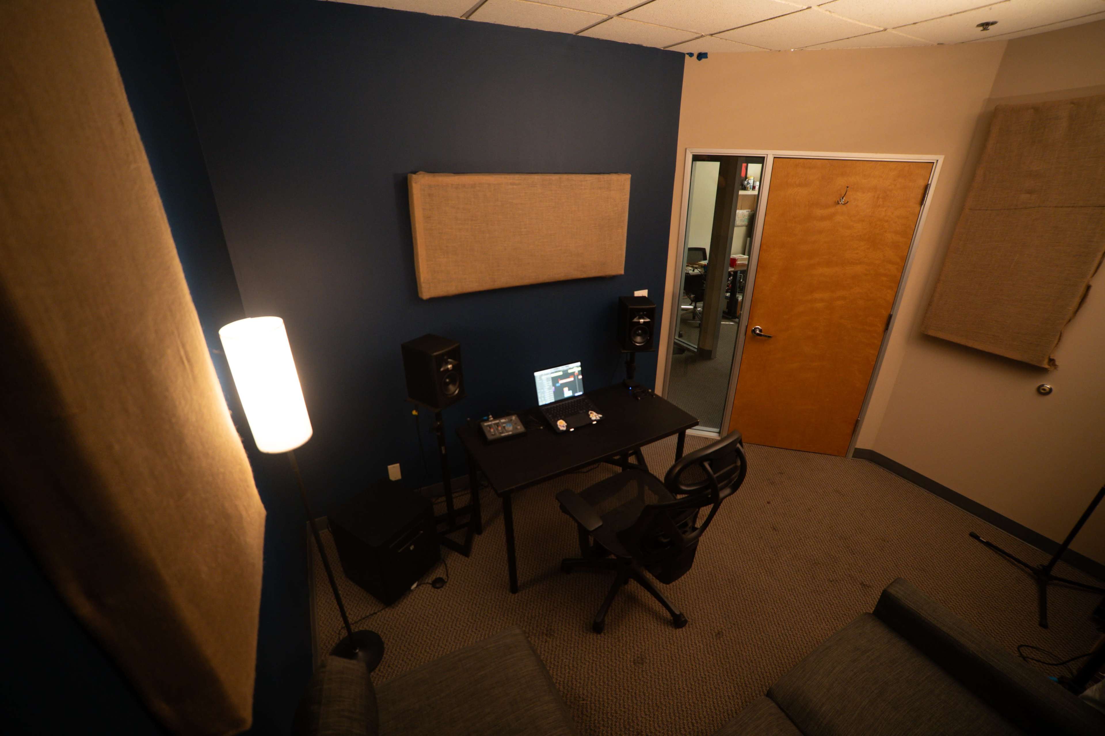 The image shows a small recording studio with a desk, computer, speakers, and soundproofing panels on the walls.