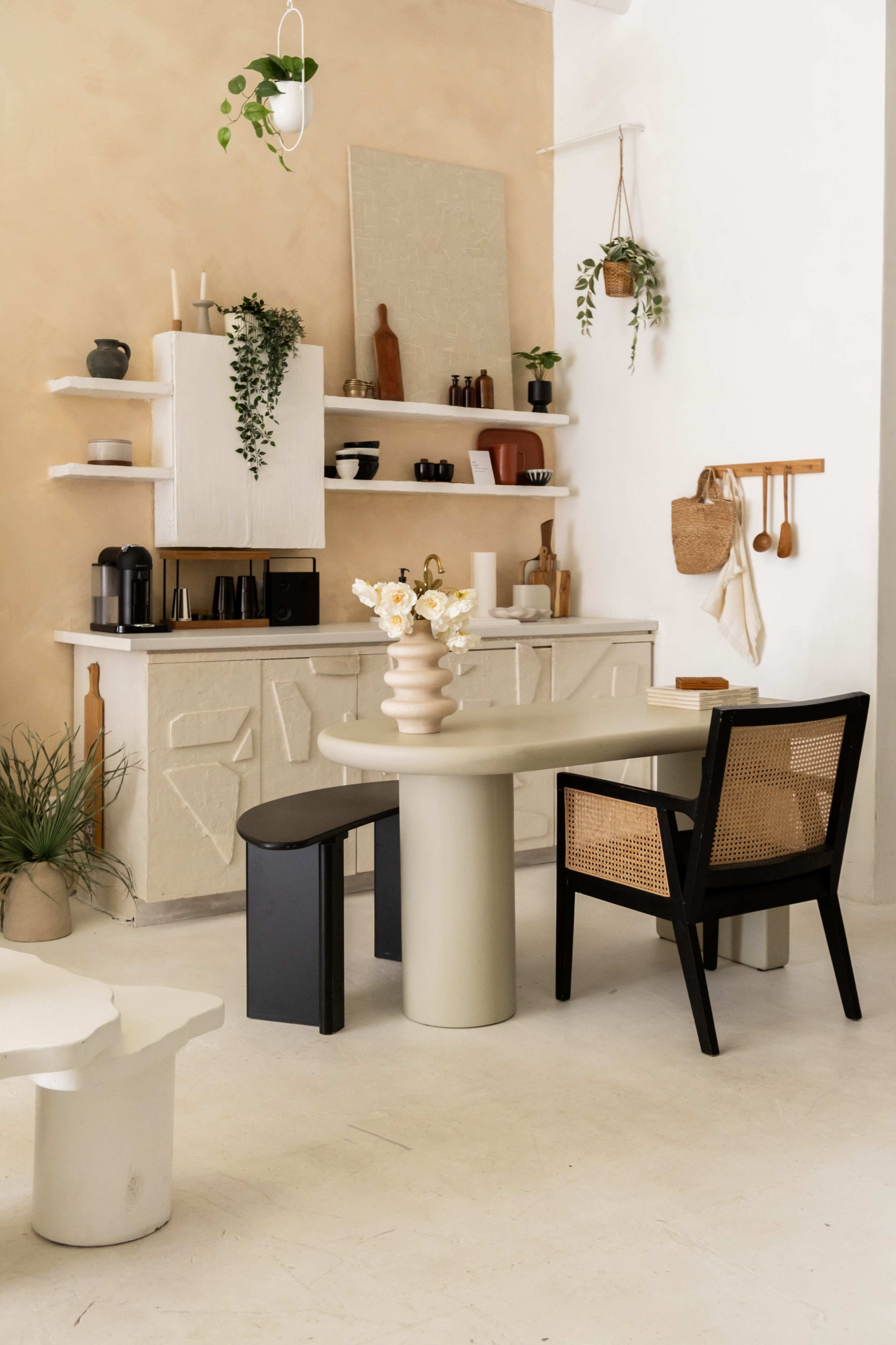The image shows a minimalist interior design featuring a light-colored dining table and black chairs, with shelves displaying various kitchen items and plants against a beige wall.