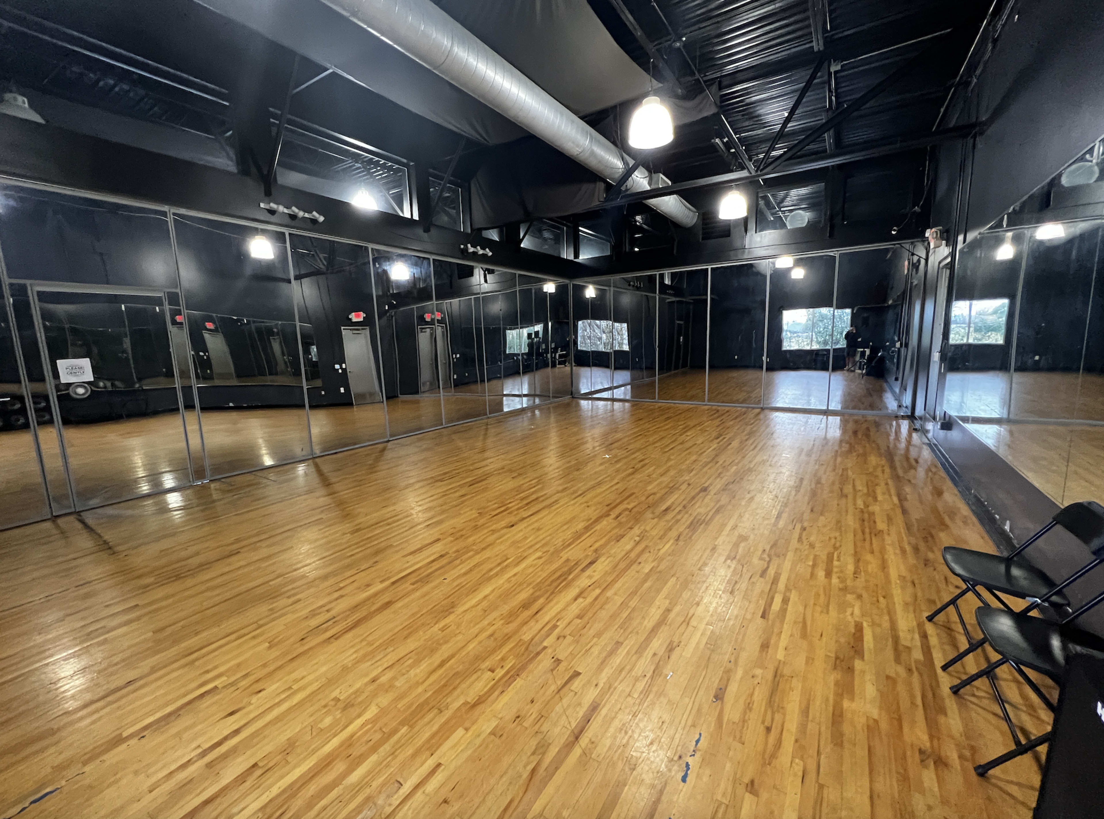 A spacious dance studio featuring wooden flooring, mirrored walls, and several folding chairs against one side.