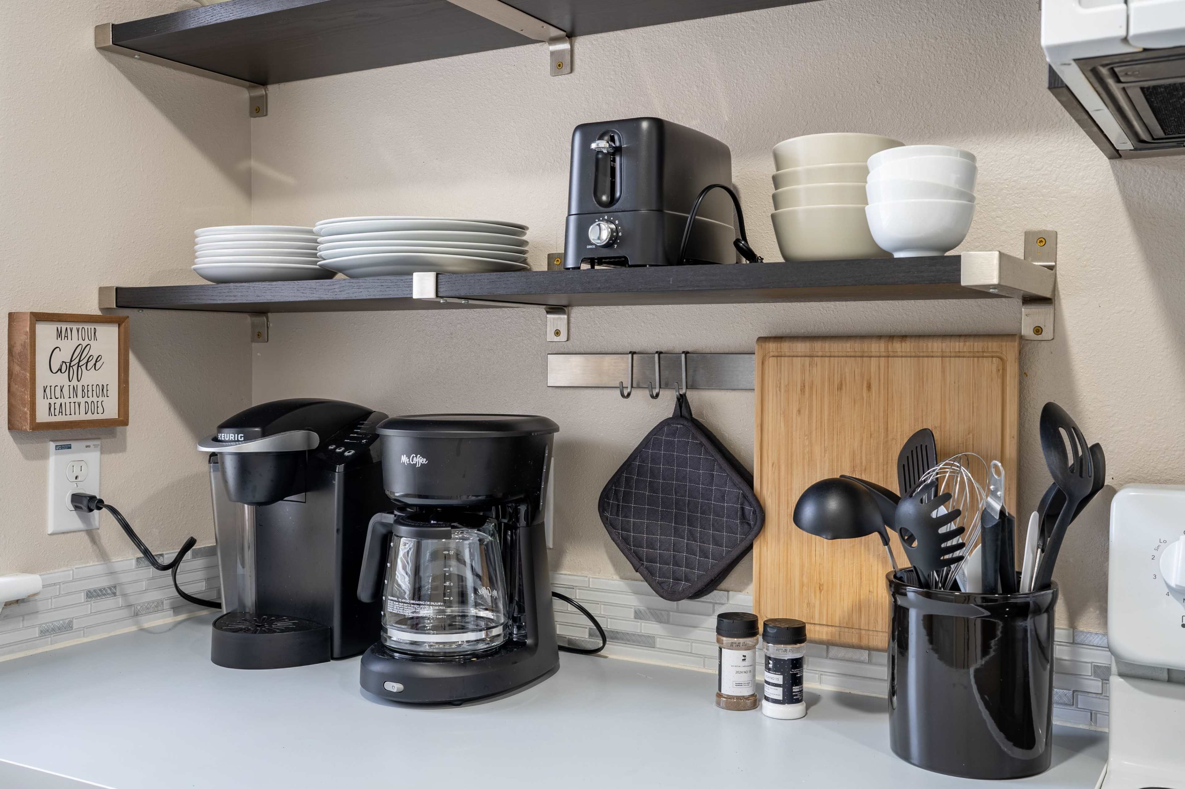 A kitchen countertop features a coffee maker, a toaster, and a coffee grinder, with neatly stacked plates and bowls on shelves above.
