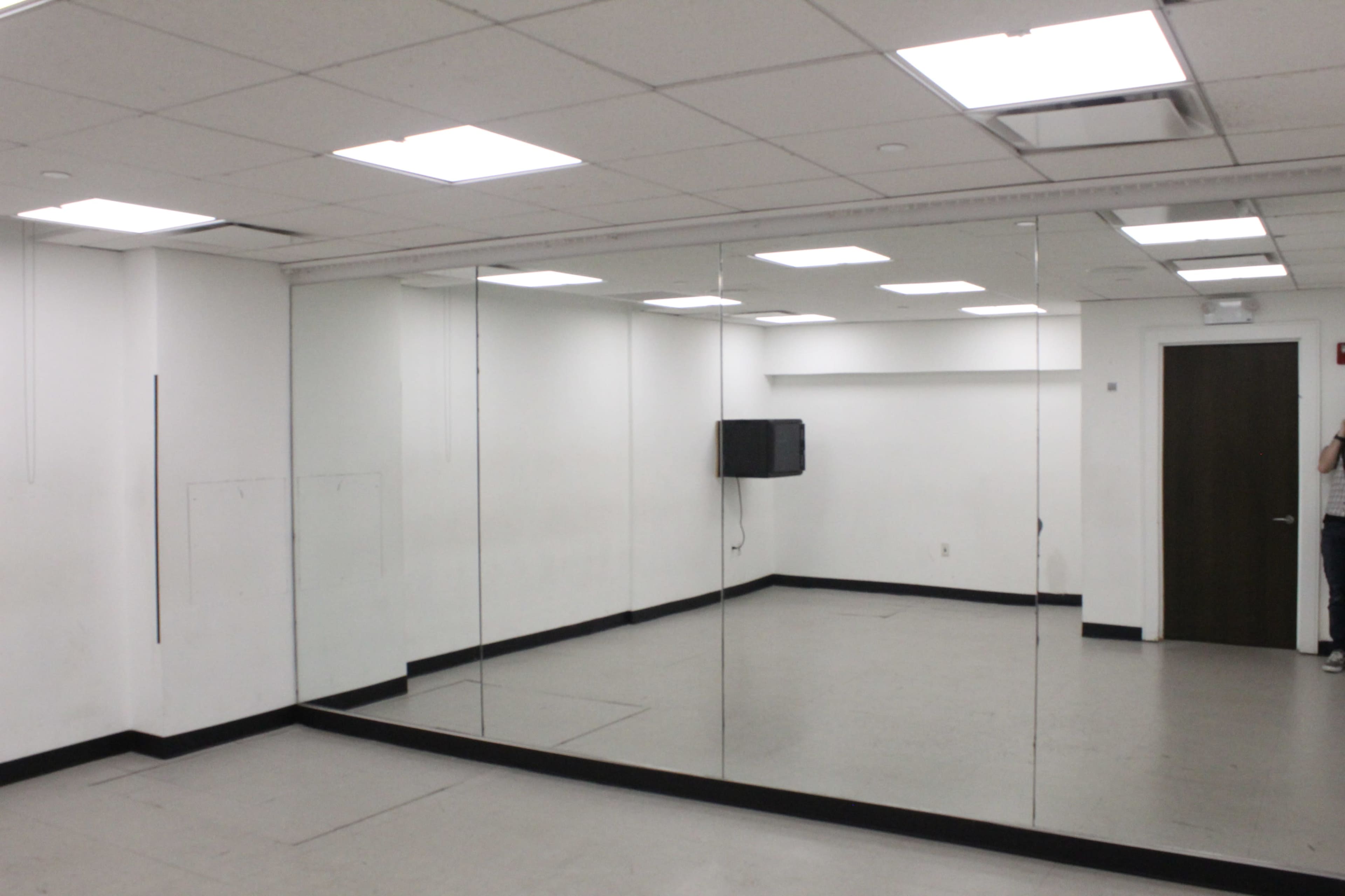 The image shows a sparsely furnished room with mirrored walls, fluorescent lighting, and a television mounted on one wall.