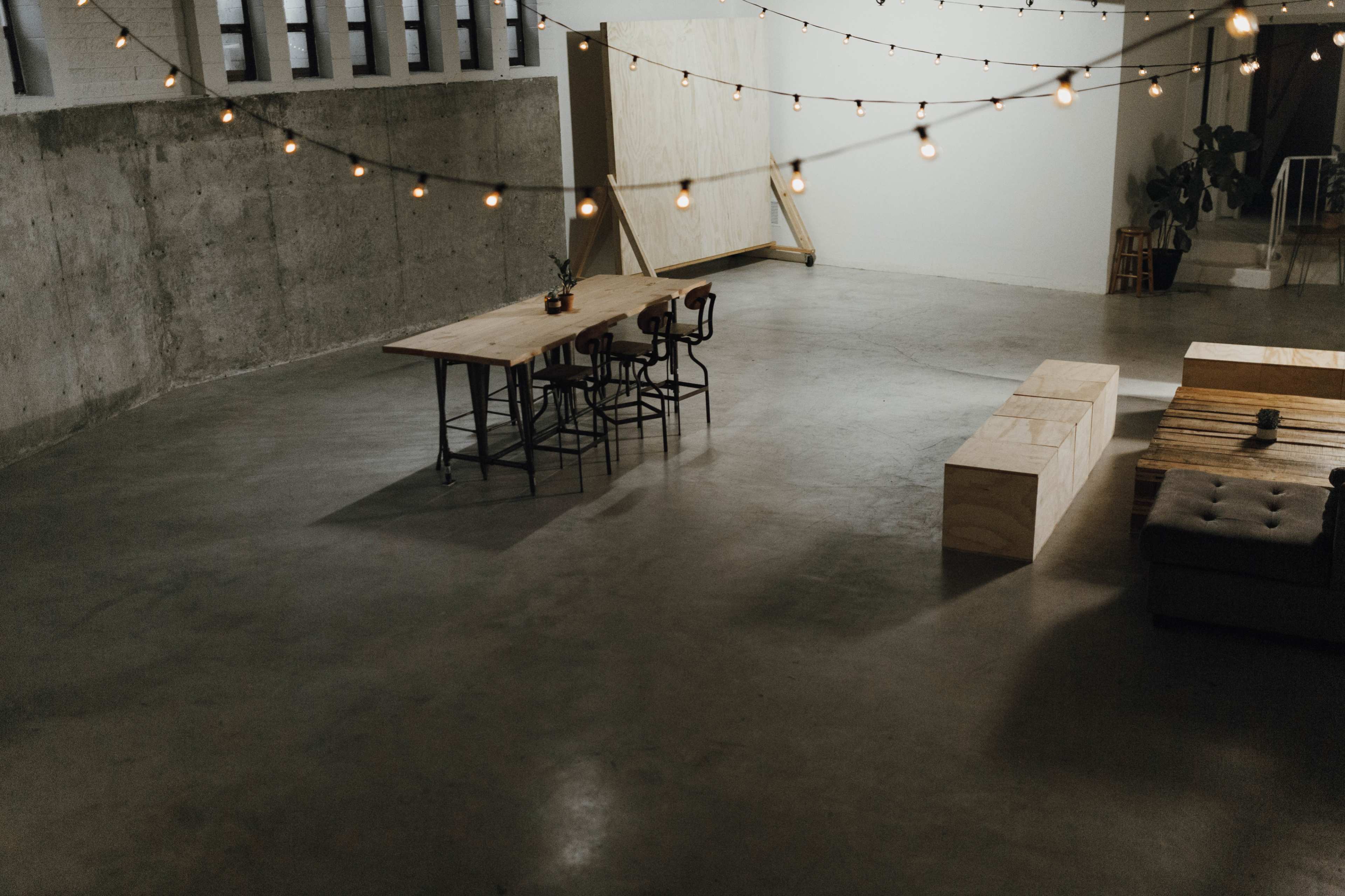 A spacious, minimalist room with a long wooden table, chairs, and string lights hanging overhead.