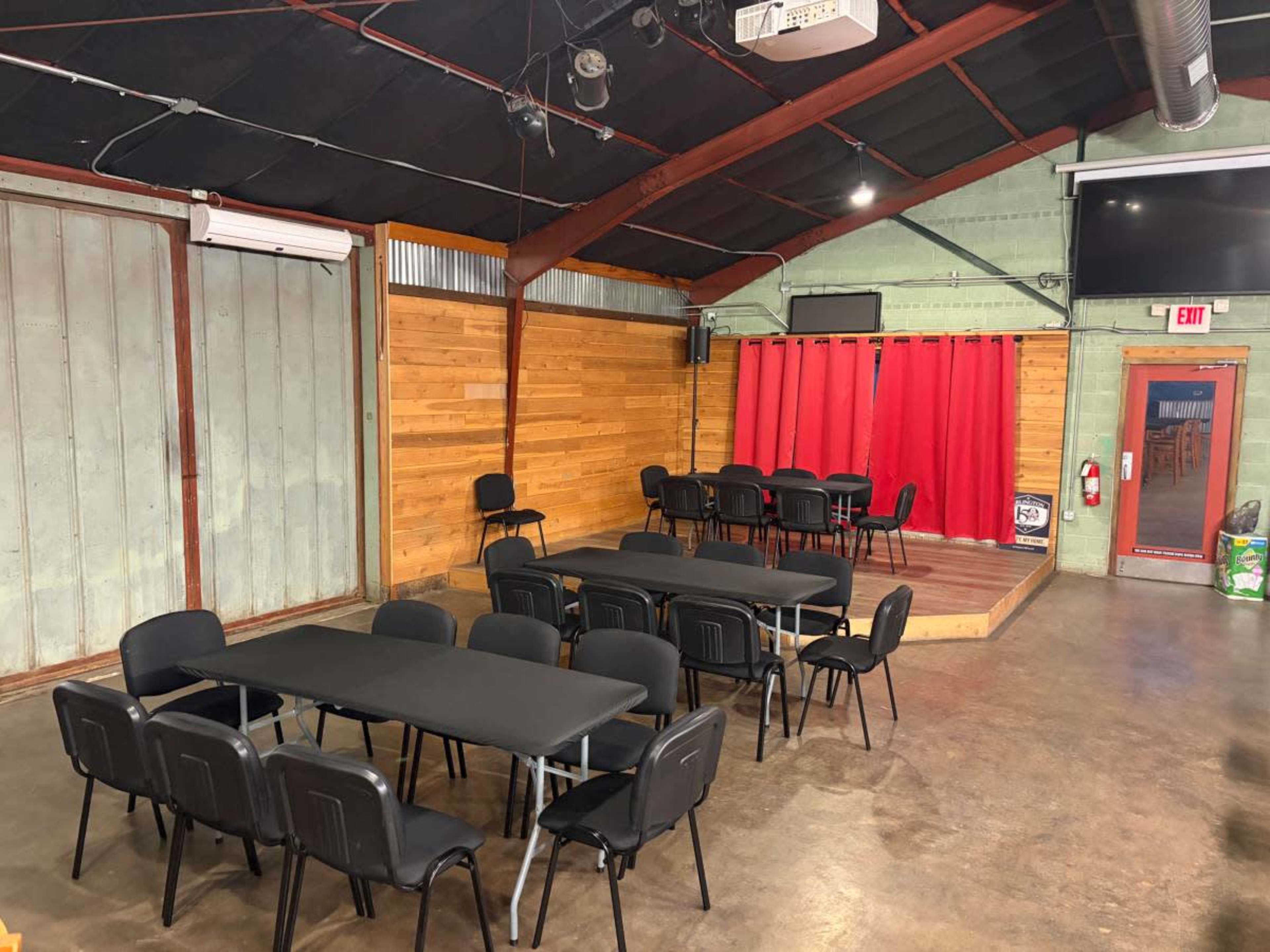 The image shows a spacious indoor venue with several black tables and chairs arranged, a stage at the back with red curtains, and wooden paneling on the walls.
