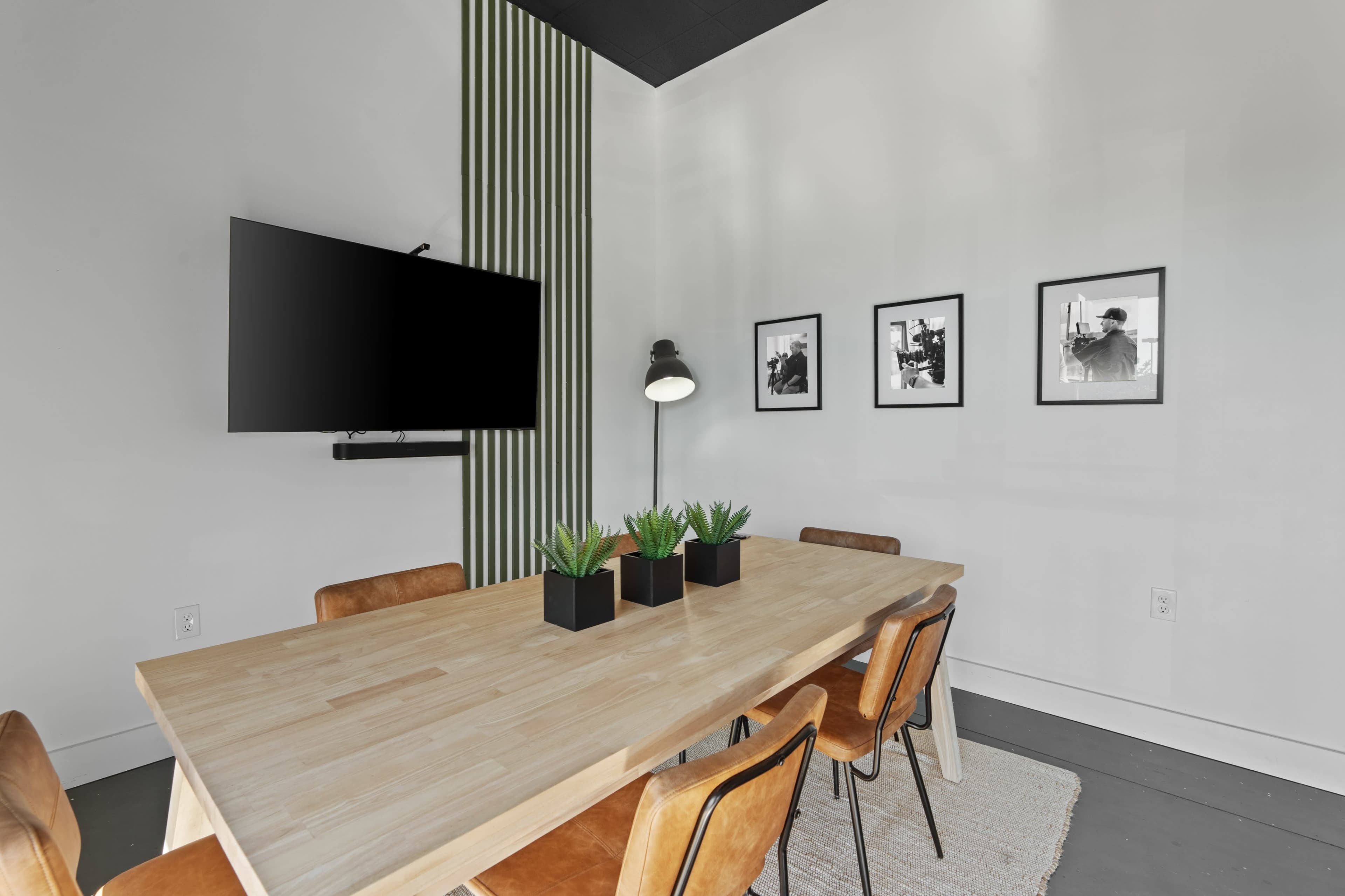 A modern conference room features a wooden table surrounded by chairs, with a mounted TV and framed black-and-white photographs on the wall.